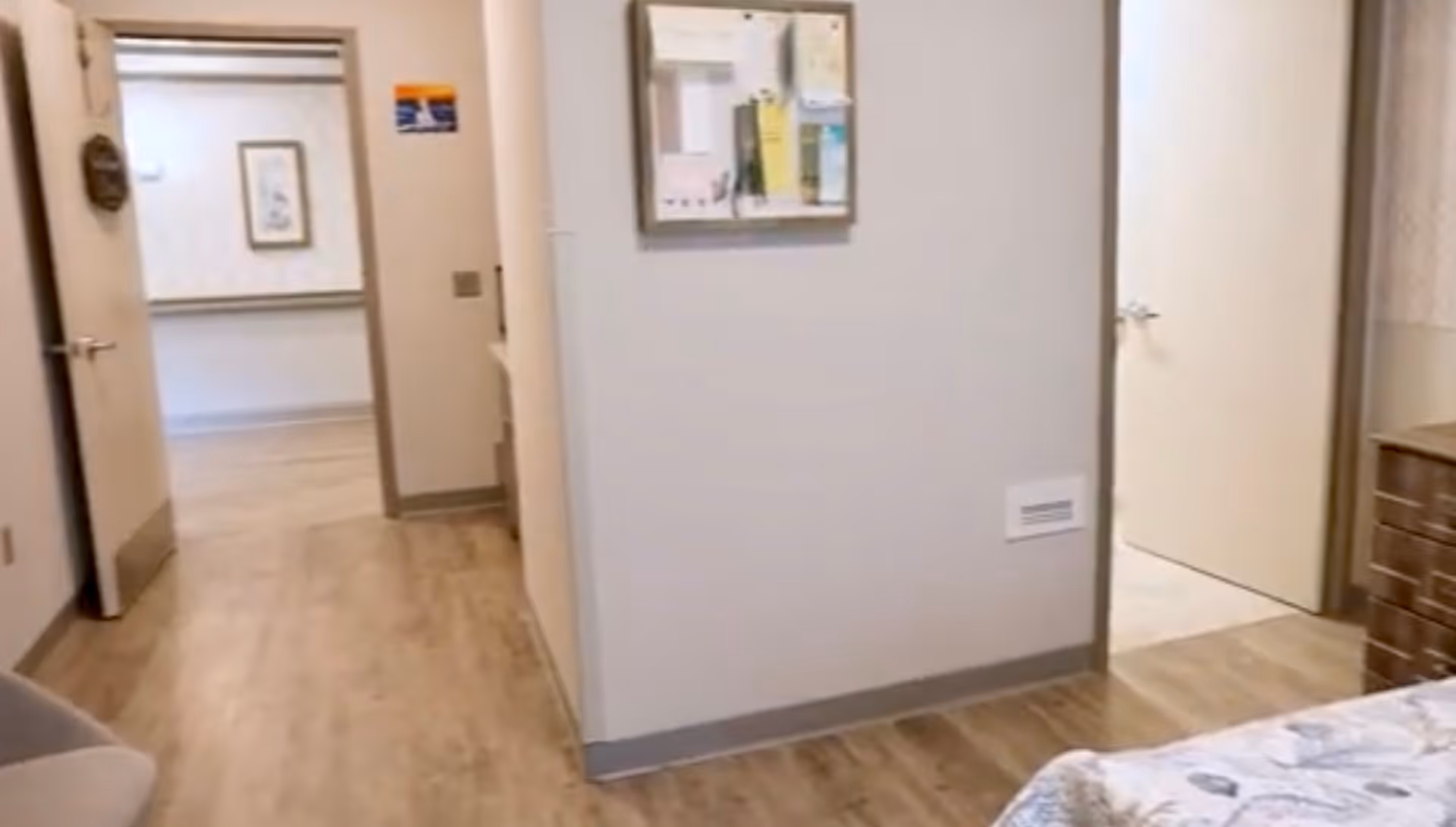 View of a hallway in a senior living facility with light wood flooring and beige walls. There are two open doors visible, one leading to another room with a framed picture on the wall and the other to a room with a bed partially visible. A bulletin board with papers is mounted on the wall in the hallway.