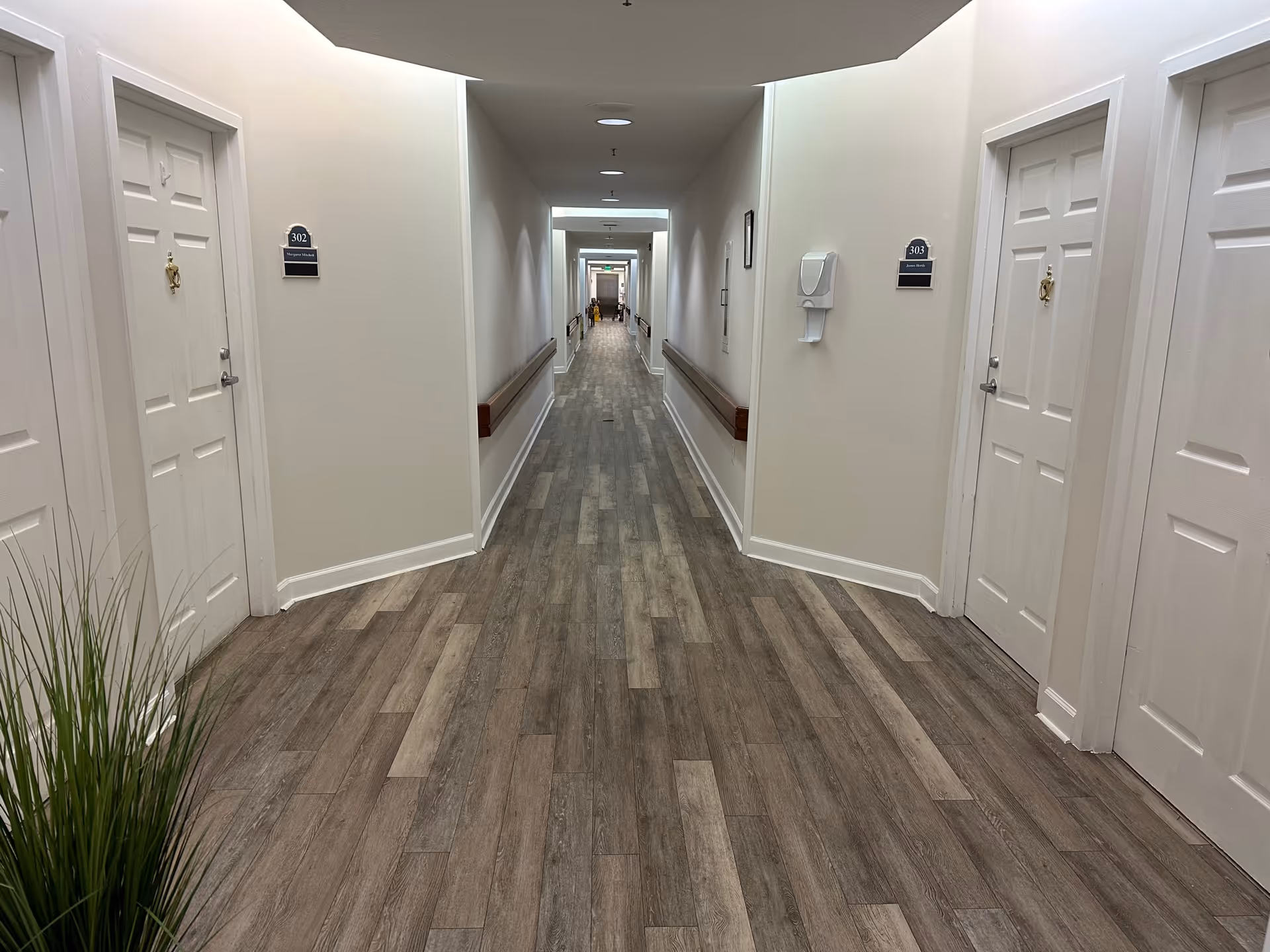 A long, well-lit hallway in a senior living facility with wood-patterned flooring, white walls, and white doors on both sides. Each door has a room number plaque and a name. Handrails run along both walls, and a hand sanitizer dispenser is mounted on the right wall. A green exit sign is visible at the far end of the hallway.