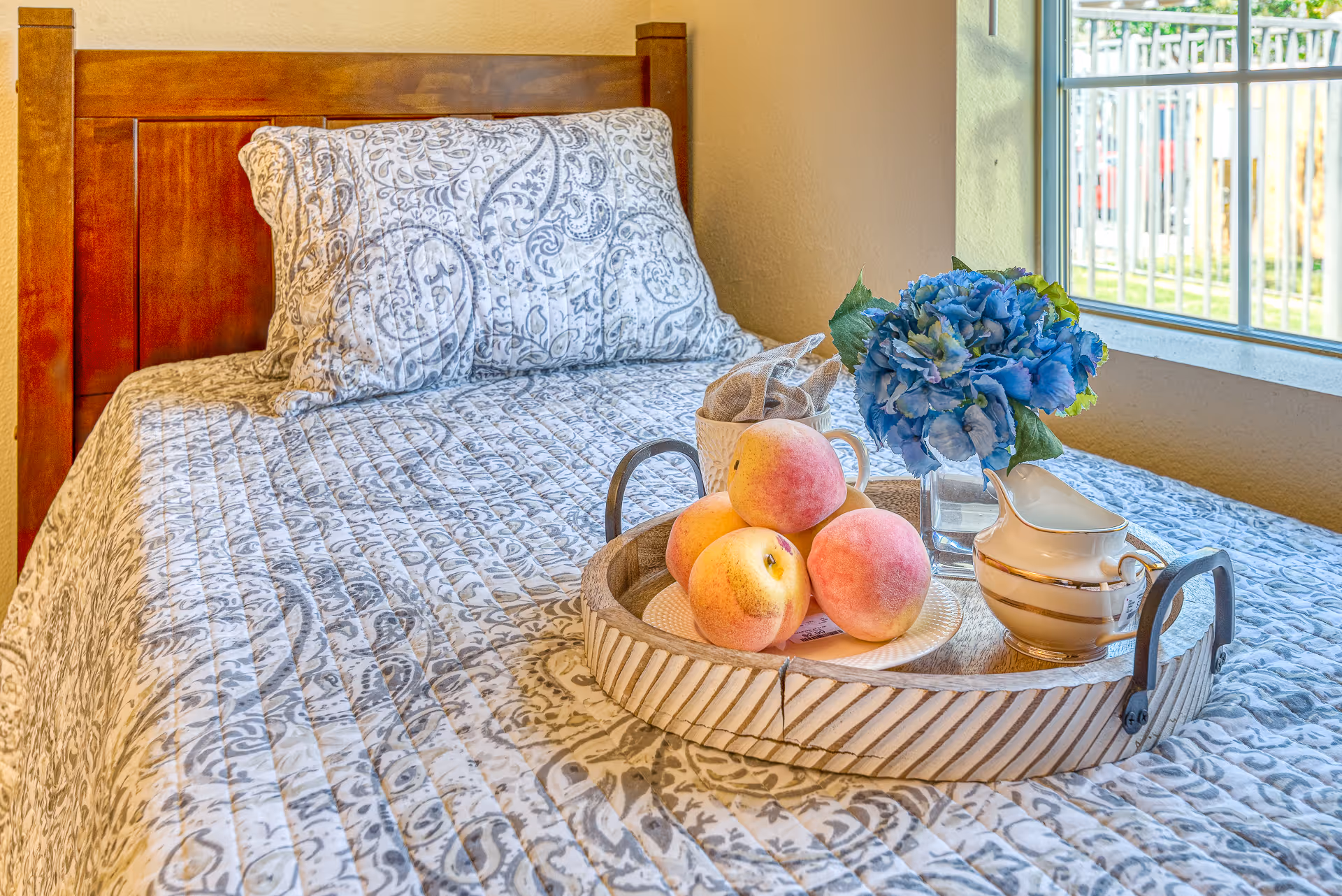 A neatly made bed with patterned bedding and a wooden headboard, a tray holding peaches, a creamer, and blue flowers by a window.