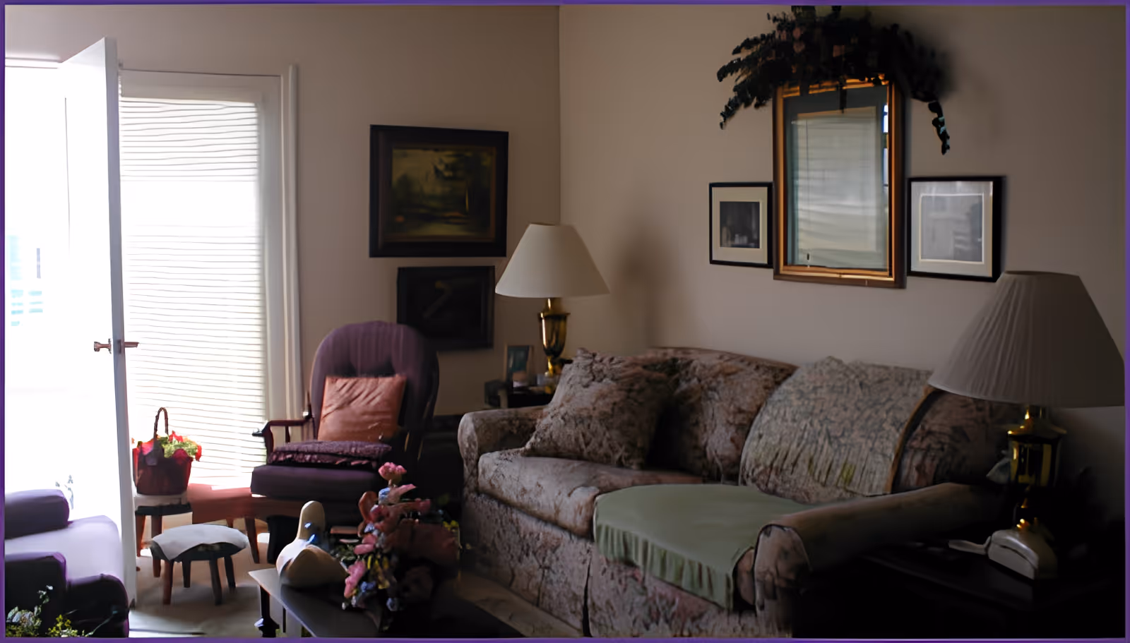 A cozy living room with a floral-patterned sofa covered partially with a green throw, a purple armchair with a pink cushion, and a small footstool. There are two table lamps with white shades on either side of the sofa. The walls are decorated with framed pictures and a mirror with greenery above it. A glass door with blinds lets in natural light, and a small table with a basket of flowers is near the armchair.