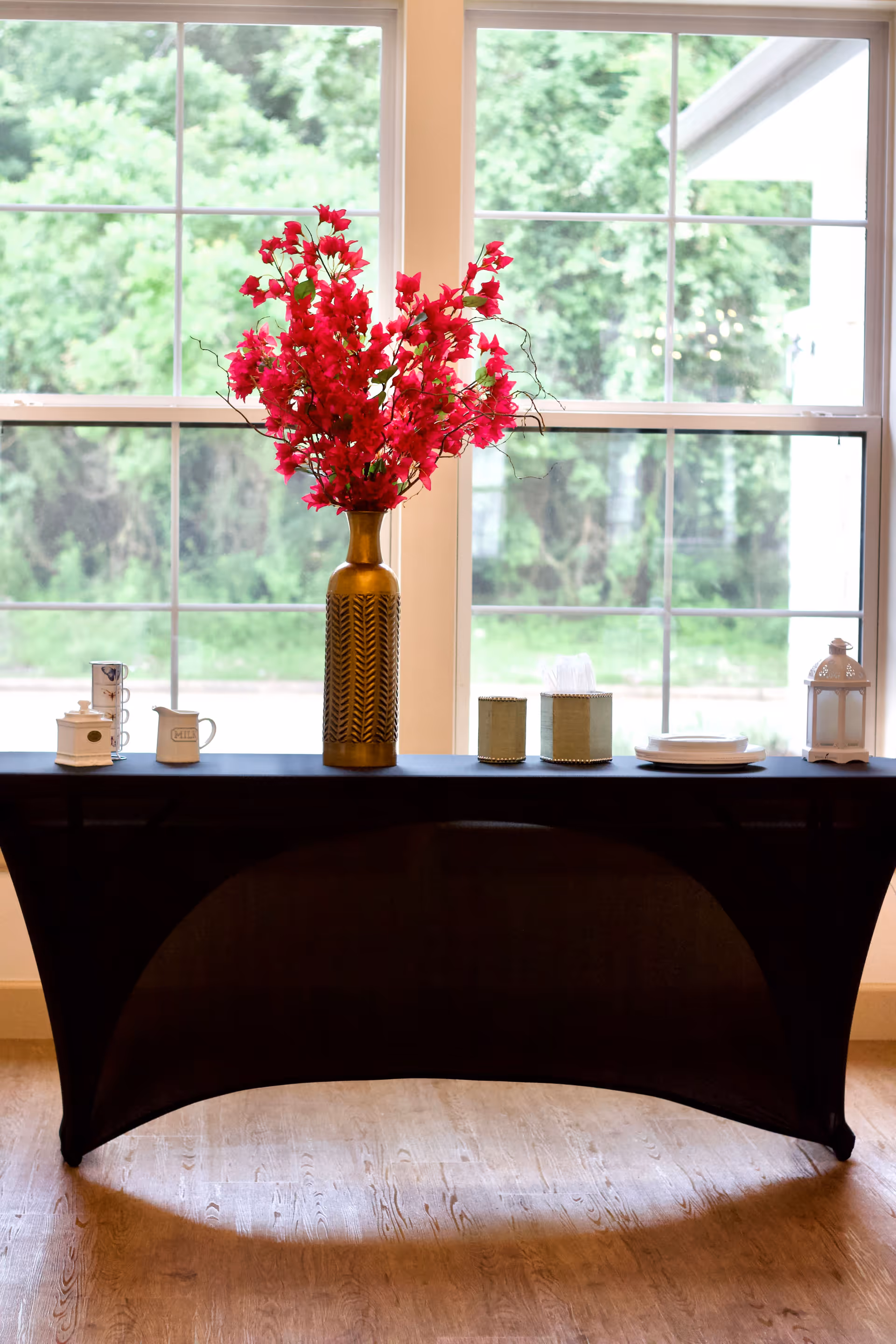 A black console table in front of a large window holding a tall gold vase of bright pink flowers and assorted decorative items.