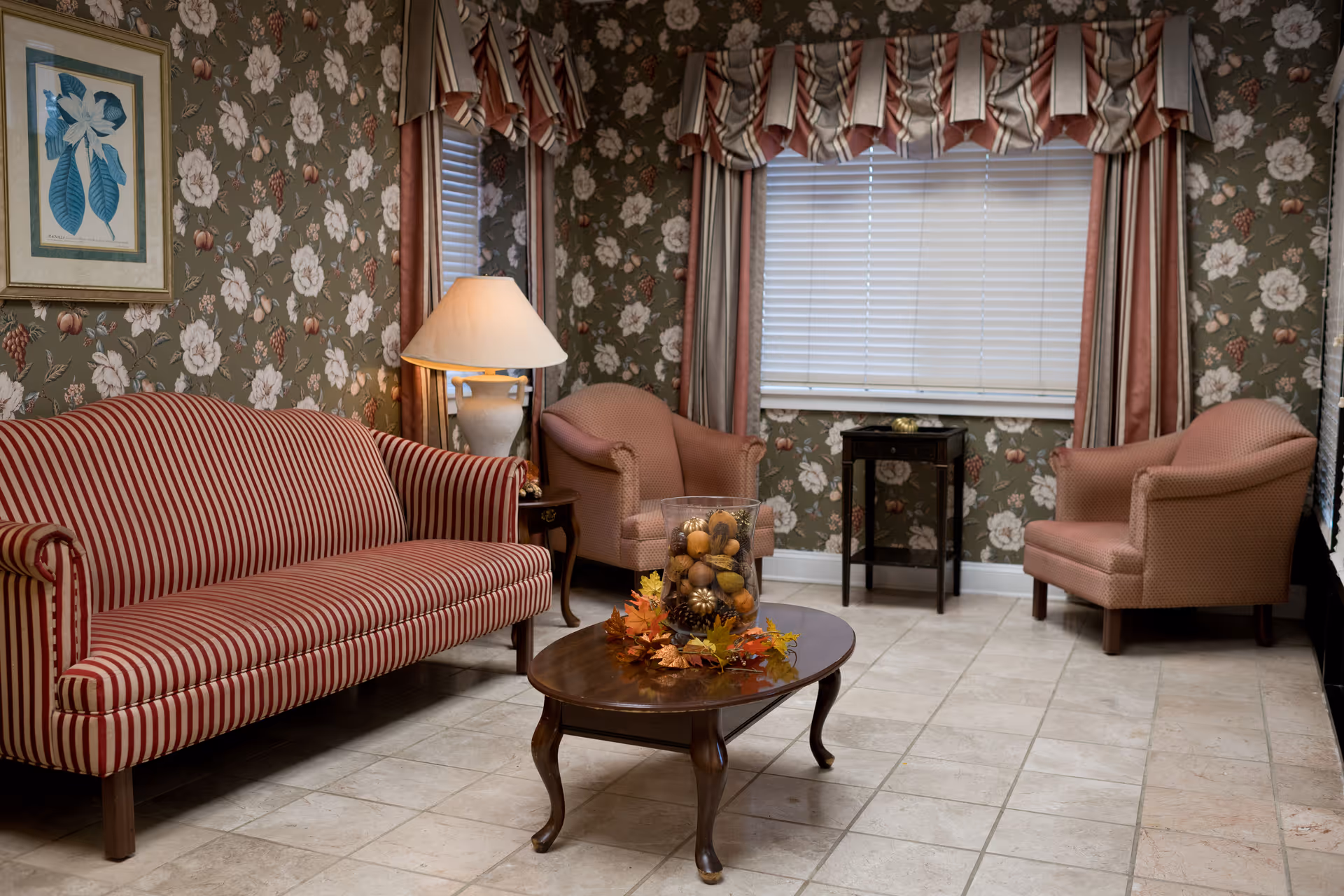 A cozy living room with floral wallpaper, a striped red and white sofa, two pink upholstered armchairs, a wooden coffee table with a decorative centerpiece of pine cones and autumn leaves, a side table with a lamp, and a window with blinds and striped curtains.