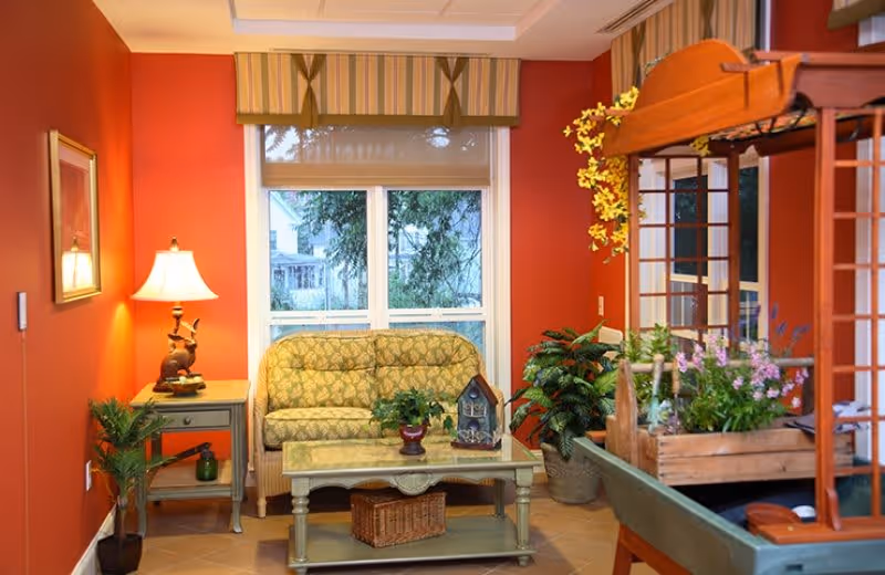A cozy living room with warm orange walls, a patterned green loveseat in front of a window with striped valance, a side table with a lamp and decorative items, a coffee table with plants and a wicker basket underneath, and a wooden plant stand with various potted flowers and greenery.