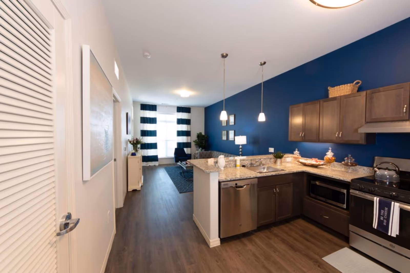 Interior view of a senior living facility apartment showing a modern kitchen with granite countertops, stainless steel appliances including a dishwasher, microwave, and stove. The kitchen has wooden cabinets and two pendant lights hanging from the ceiling. Beyond the kitchen is a living area with a blue accent wall, a couch, a chair, a lamp, and a window with blue and white striped curtains.