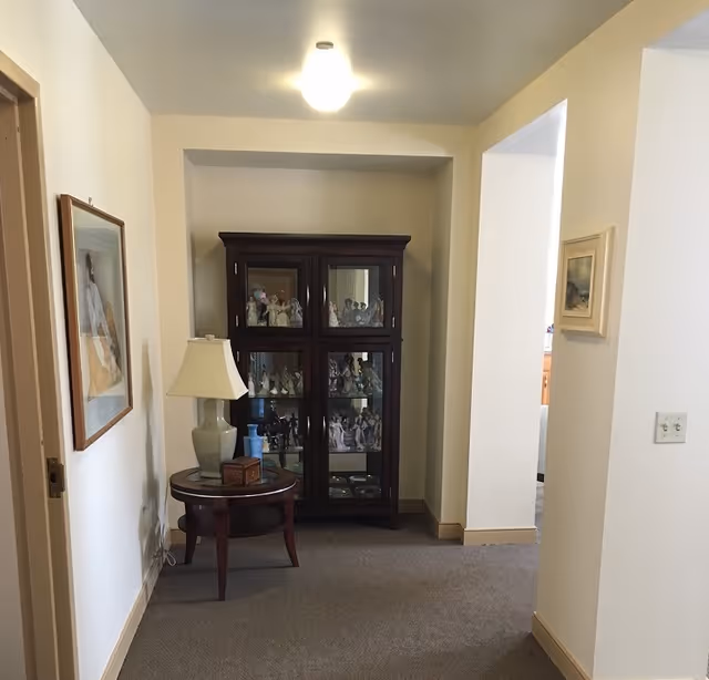 A hallway area in a retirement community with beige walls and carpeted floor. At the end of the hallway is a dark wooden display cabinet filled with figurines. To the left of the cabinet is a round wooden side table with a lamp, a small box, and a bottle of hand sanitizer. There are framed pictures hanging on the walls and a ceiling light fixture above.