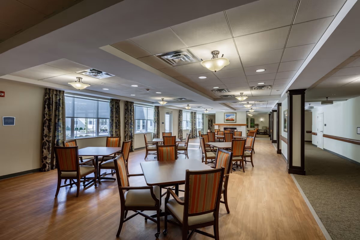 A spacious dining room with multiple wooden tables and chairs with striped upholstery. Large windows with patterned curtains allow natural light to fill the room. The ceiling has recessed lighting and decorative light fixtures. The floor is a combination of wood and carpet, and there is a hallway visible on the right side.