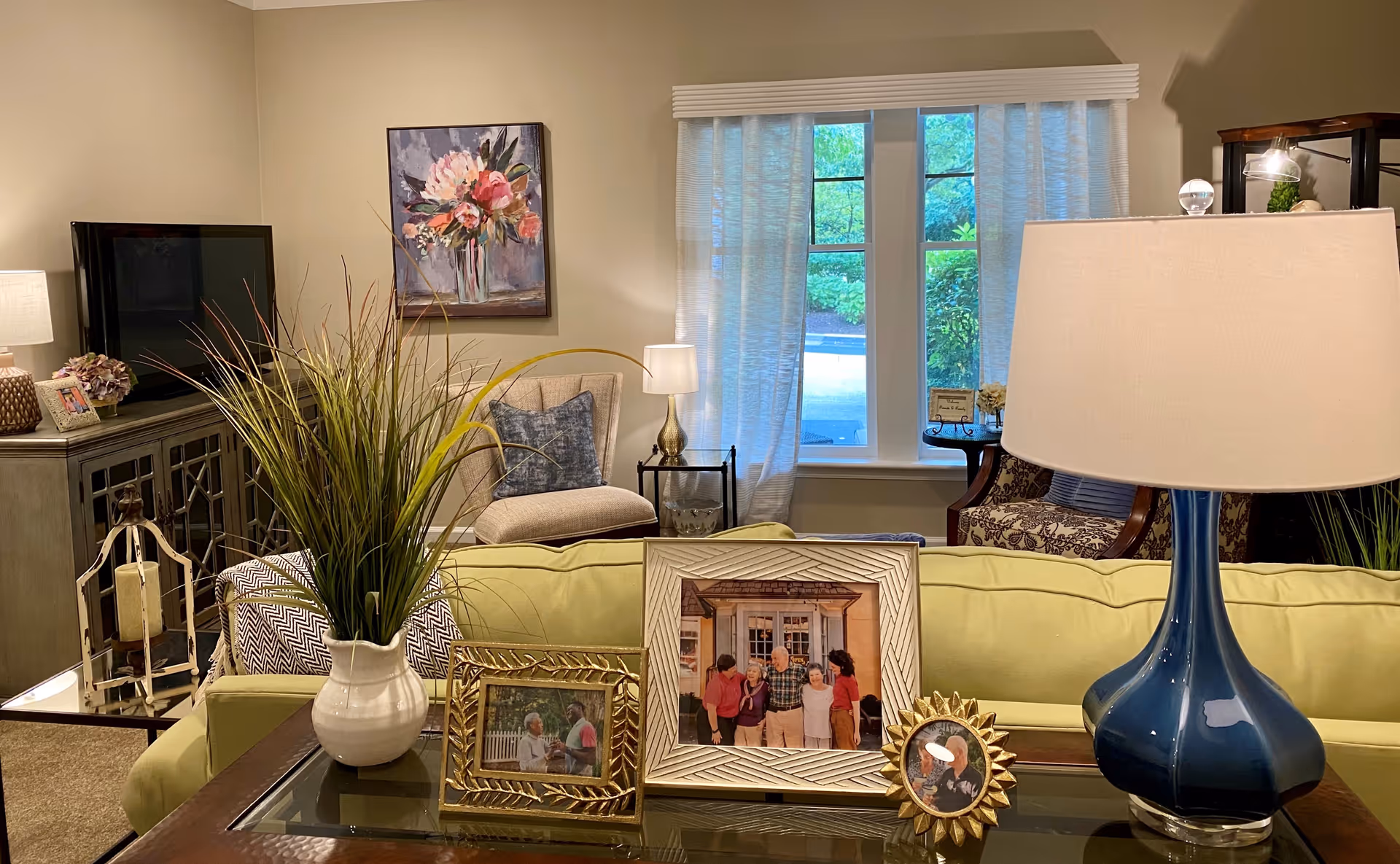 A cozy living room with a green sofa, a blue table lamp, and three framed family photos on a glass coffee table. Behind the sofa, there is a beige armchair with a blue pillow, a side table with a lamp, and a window with sheer white curtains showing greenery outside. A TV sits on a cabinet to the left, and a floral painting hangs on the wall above the armchair.