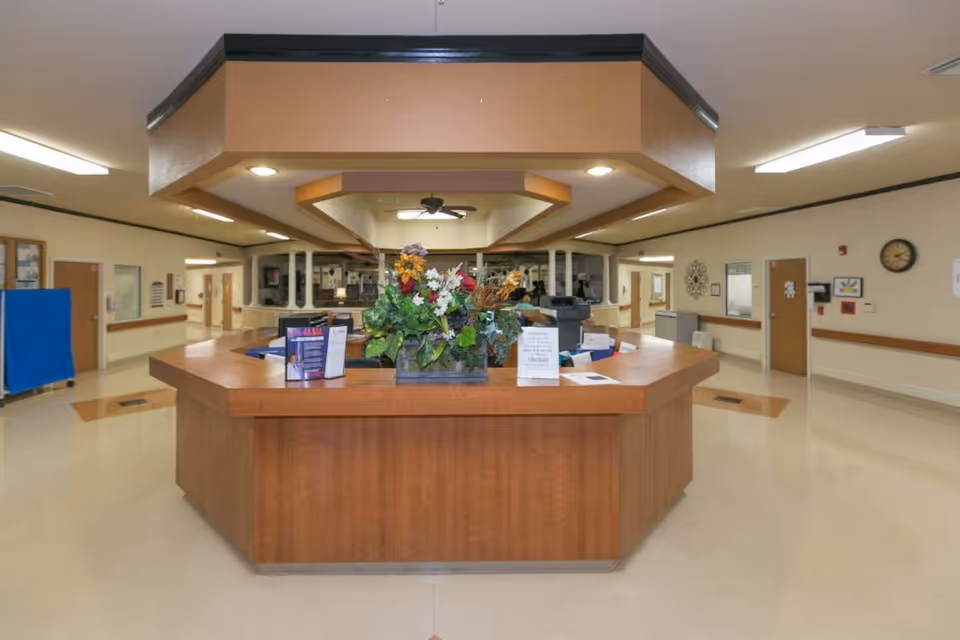 A spacious reception or nurse station area in a healthcare facility with a large wooden octagonal desk in the center. The desk has a floral arrangement and informational pamphlets on top. The surrounding area has beige walls, multiple doors, a clock on the wall, and overhead lighting.