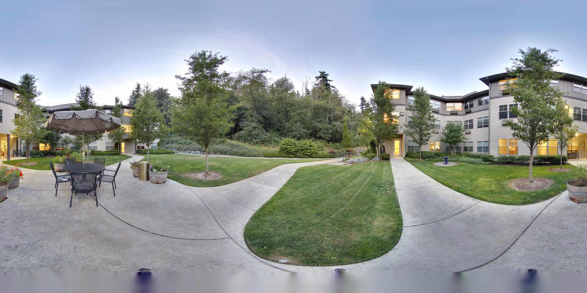 Outdoor courtyard area of a senior living facility with concrete walkways, green grass, trees, and a patio table with chairs and an umbrella. The surrounding building has multiple windows and is three stories tall. The sky is clear and it appears to be early evening.