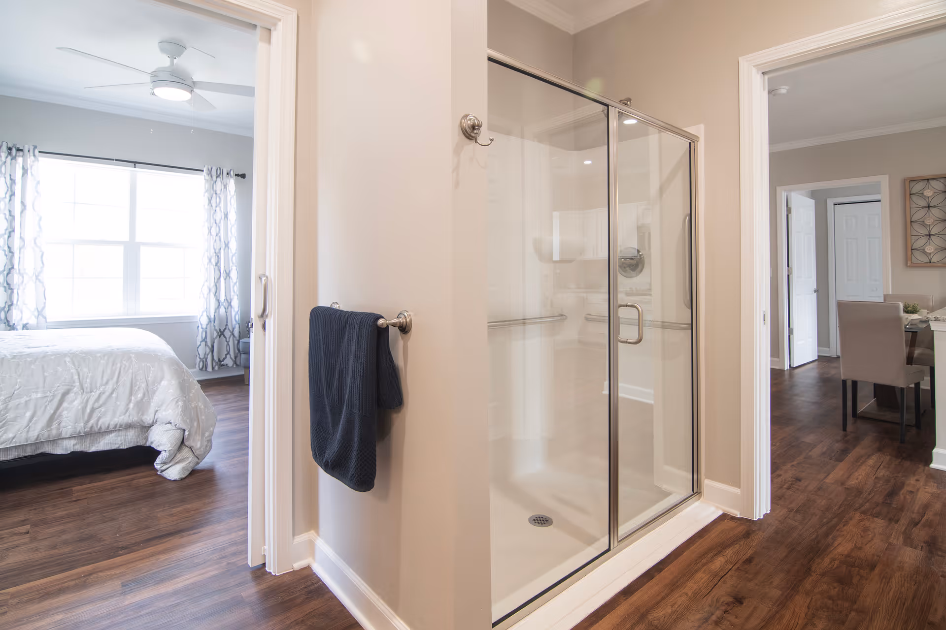 Interior view showing a glass-enclosed shower with a dark towel hanging on a wall-mounted towel rack. To the left, there is a bedroom with a bed, white bedding, and a large window with patterned curtains. To the right, a dining area with a table and chairs is visible. The flooring is dark wood throughout.