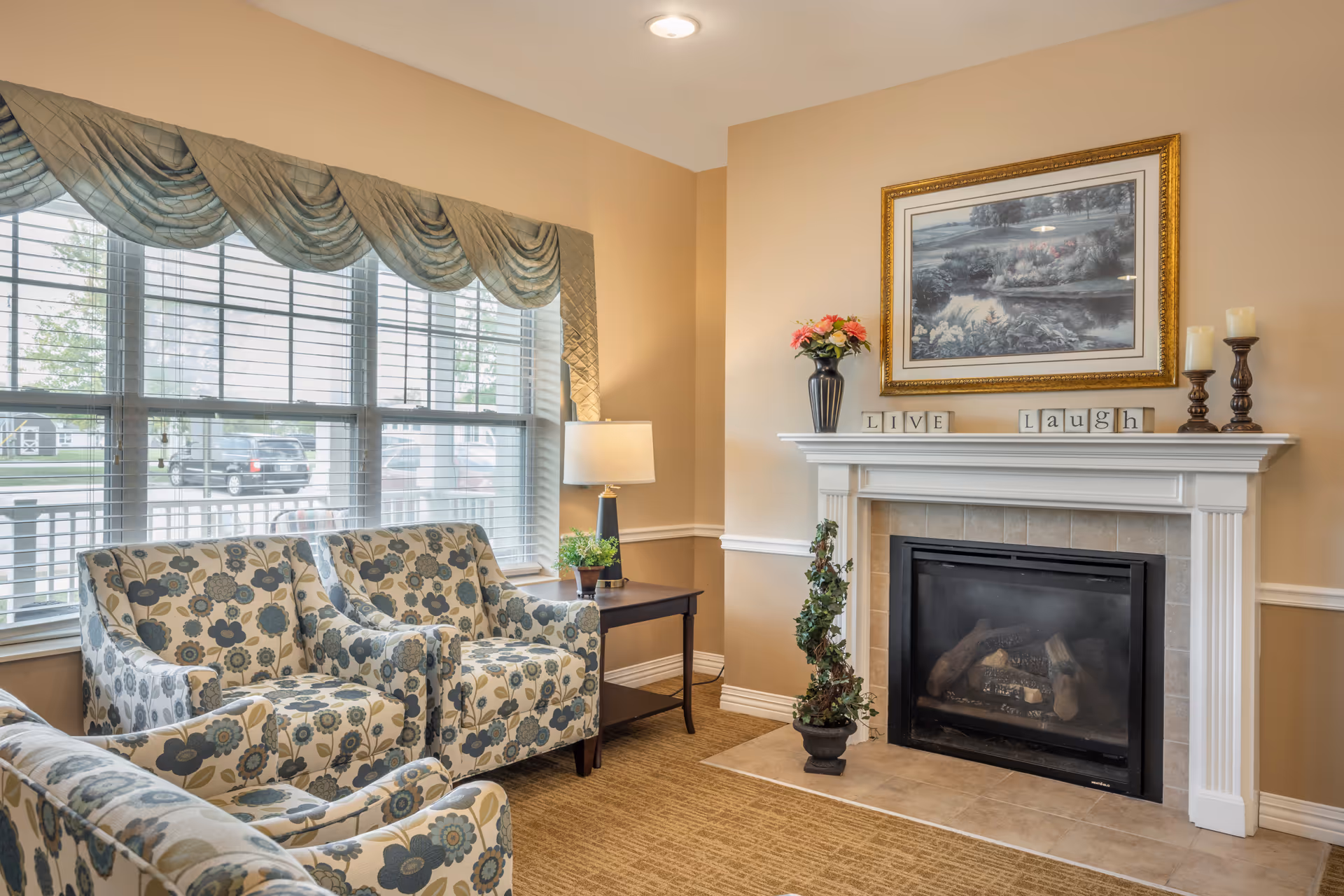 A cozy living room area with three floral patterned armchairs arranged near a window with blinds and a decorative valance. A side table with a lamp and a small plant is placed between the chairs. There is a fireplace with a white mantel decorated with a vase of flowers, candles, and blocks spelling out 'LIVE' and 'Laugh'. Above the mantel hangs a framed landscape painting.