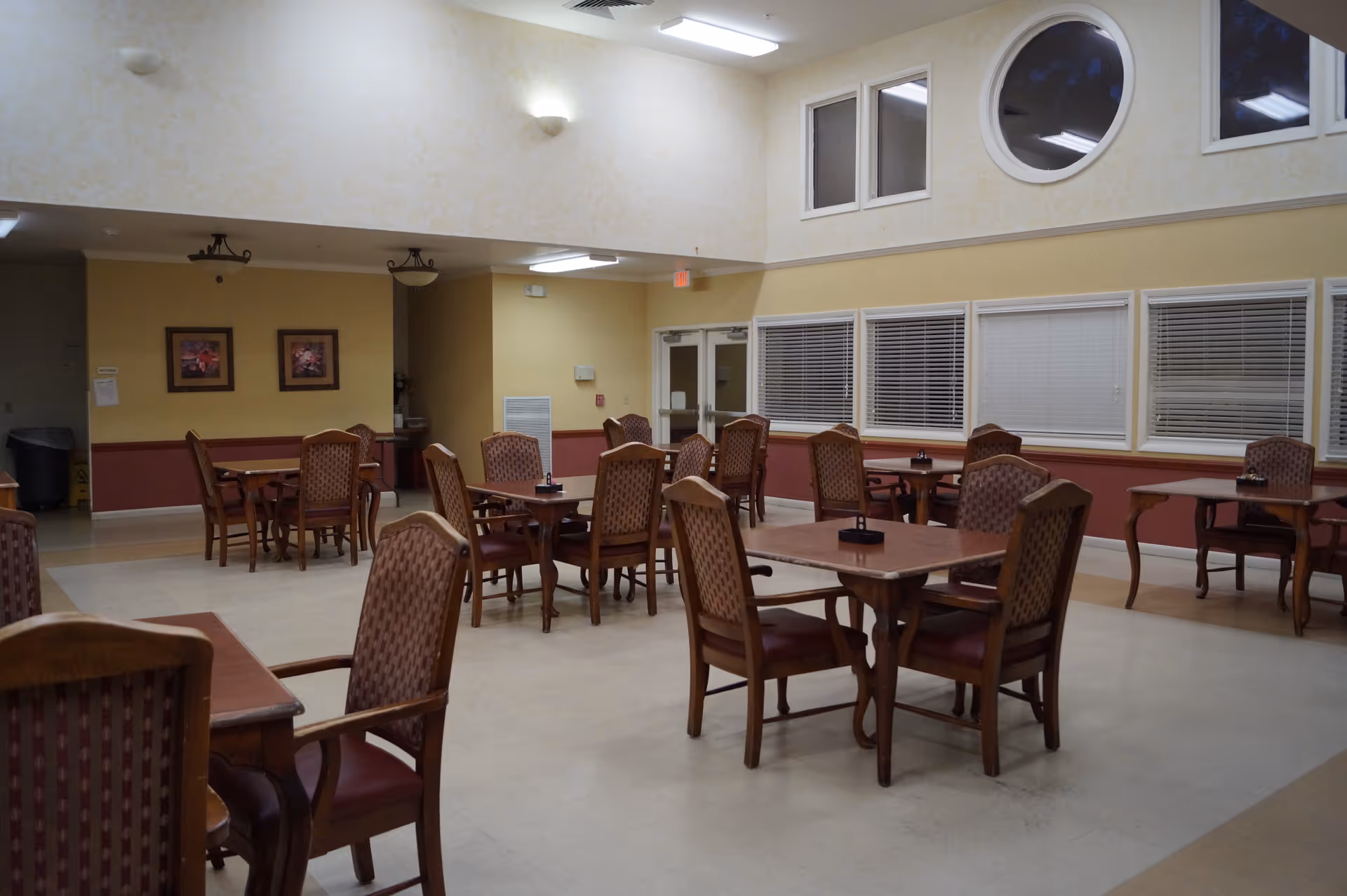 A dining room in The Stratford facility with multiple wooden tables and cushioned chairs arranged neatly. The room has high ceilings with a round window and several rectangular windows with blinds. The walls are painted in light yellow and beige tones, and there are framed pictures on one wall. The room is well-lit with ceiling lights.