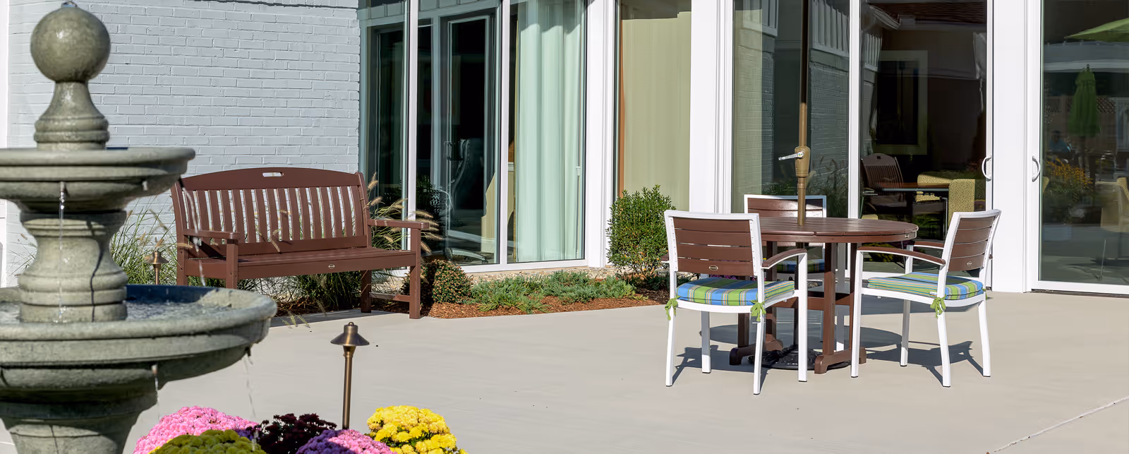 Outdoor patio area with a stone water fountain in the foreground, a wooden bench, and a round table with three chairs that have blue and green striped cushions. The patio is adjacent to a building with large glass doors and windows.