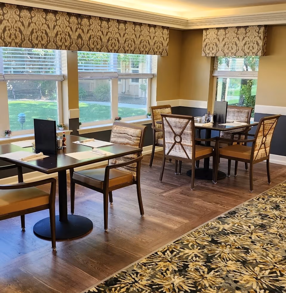A dining area in a senior living facility with two tables, each surrounded by four cushioned chairs. The room has large windows with patterned valances, allowing natural light to brighten the space. The floor is wooden with a patterned carpet along one side. Tables are set with placemats, napkins, and condiments.