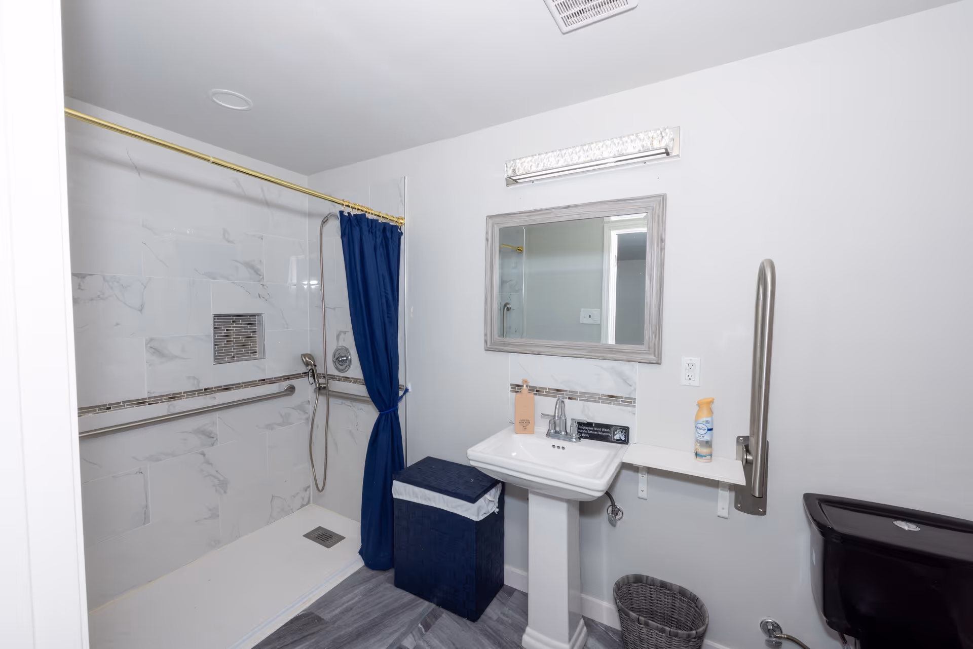 A clean, accessible bathroom featuring a walk-in shower with a blue curtain, grab bars, a pedestal sink with a mirror above it, a small shelf with a spray bottle, a dark-colored toilet, a laundry basket, and a waste bin on a gray tiled floor.