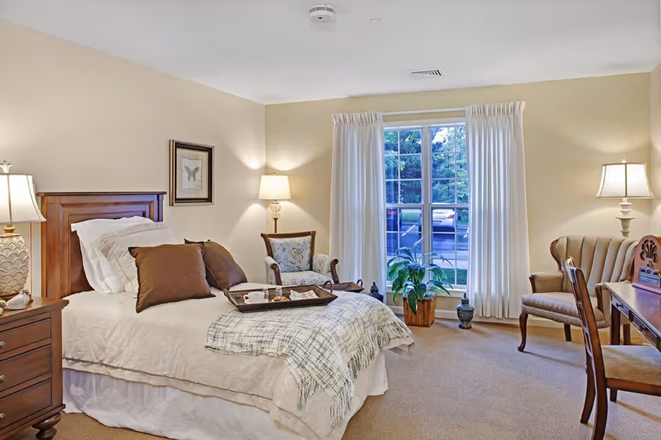 A cozy bedroom in a senior living facility with a neatly made bed featuring brown pillows and a plaid throw blanket. There is a wooden nightstand with a lamp on the left side of the bed, two upholstered chairs near a large window with white curtains, a potted plant on the floor, and a wooden desk with a chair and a lamp on the right side of the room.