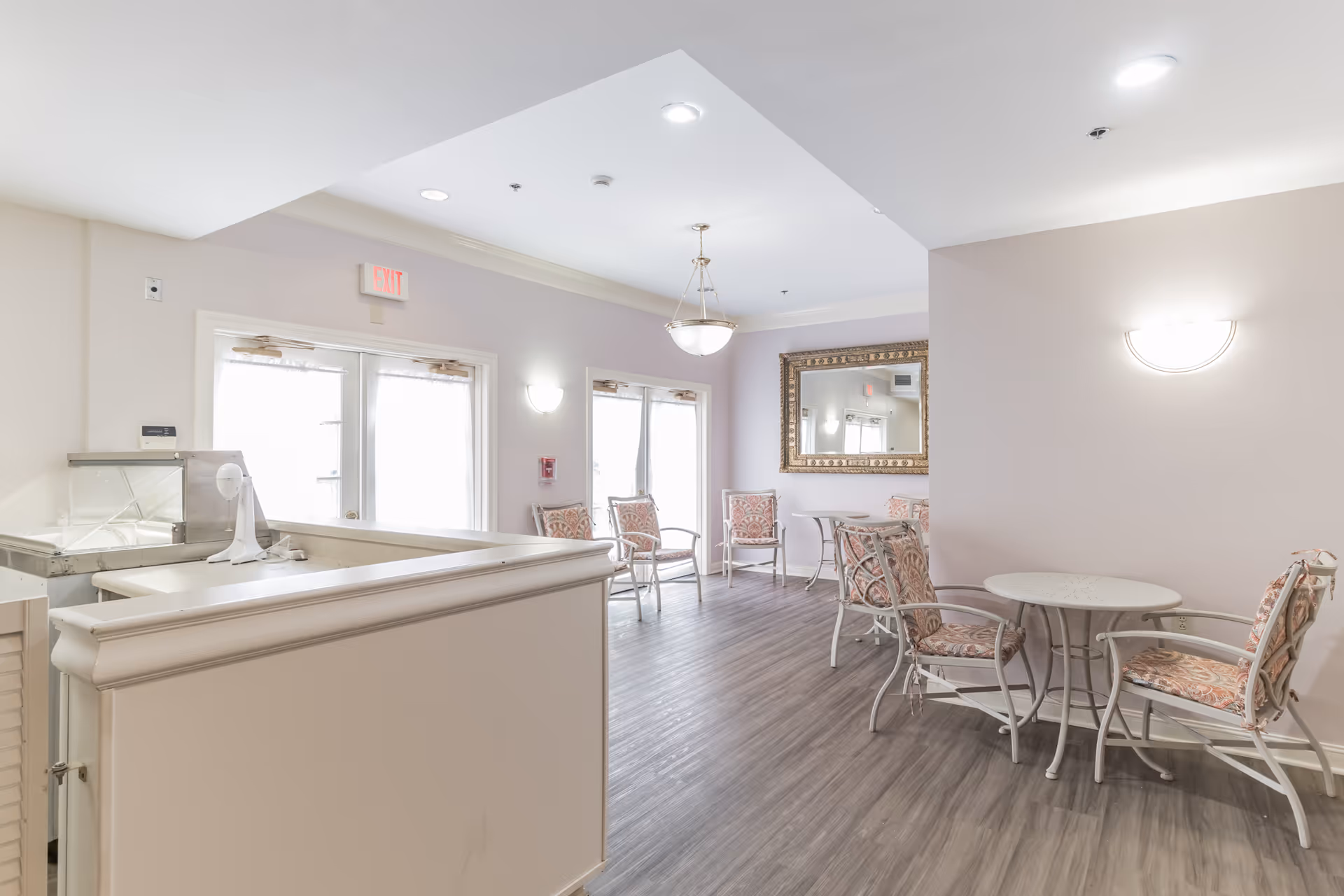 A bright and clean common area in The Rutherford Assisted Living featuring several cushioned chairs with patterned upholstery arranged around small round tables. There is a large ornate mirror on the wall, a hanging light fixture, and double doors with sheer curtains allowing natural light to enter. The floor is wood-style laminate, and the walls are painted a soft pastel color.