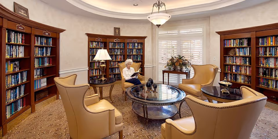 A cozy senior living library room with wooden bookshelves filled with books, beige leather armchairs arranged around a glass coffee table, a table lamp, and a window with white shutters. An elderly woman is sitting in one of the armchairs reading a book.