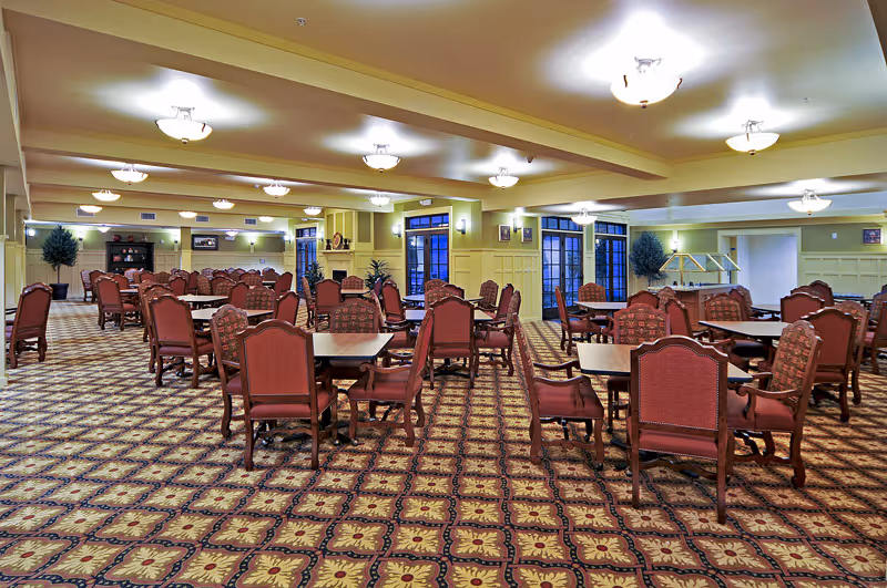 Large dining room with many square tables and red upholstered chairs arranged on a patterned carpet under ceiling light fixtures and windows.