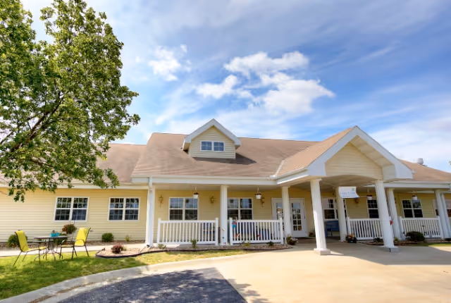 Exterior view of a single-story yellow senior living facility building with a covered entrance, white railings, and a small outdoor seating area with a table and chairs on the lawn. The sky is partly cloudy and there is a tree on the left side.