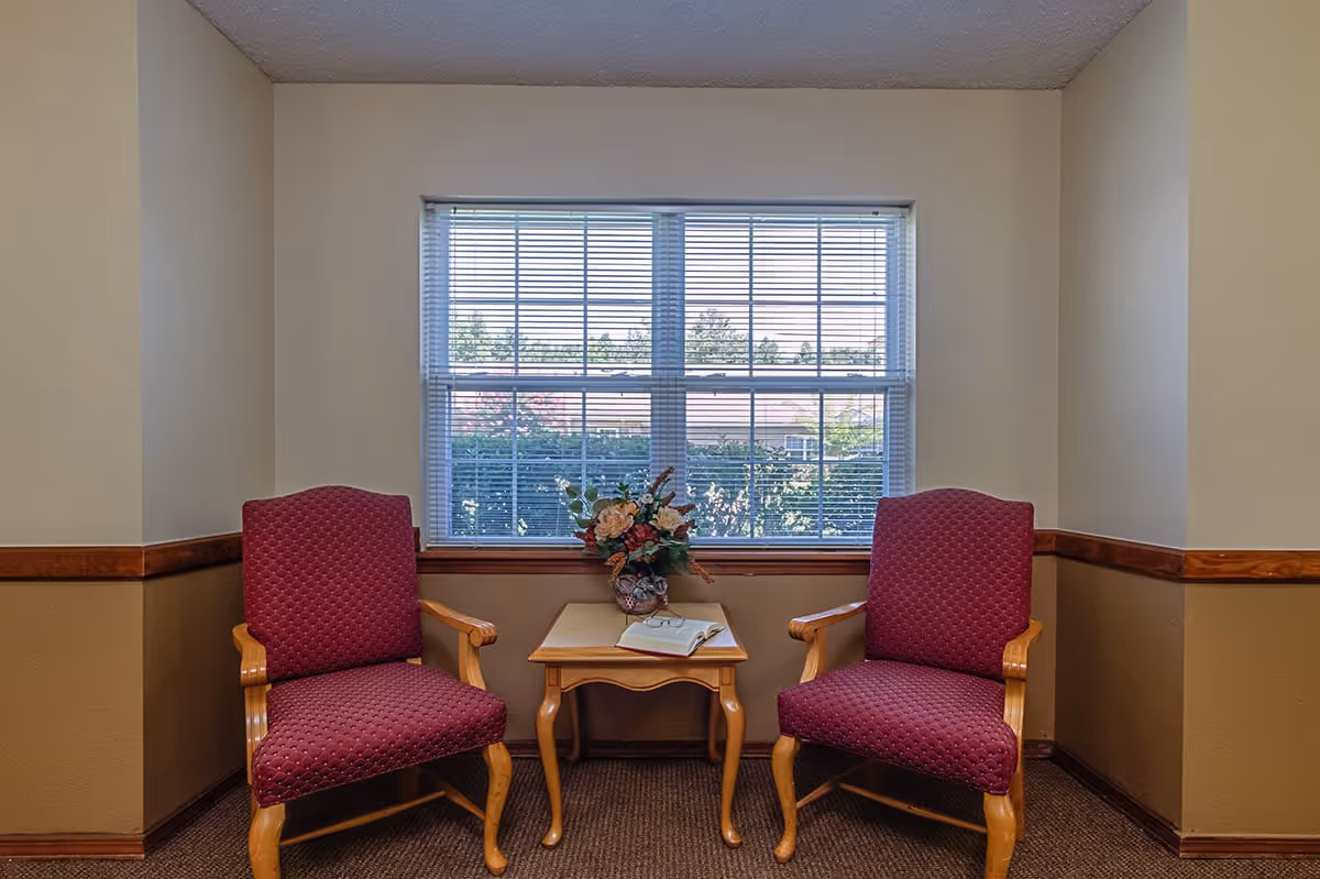 Two red upholstered armchairs with wooden arms and legs are placed on either side of a small wooden table with curved legs. On the table is a floral arrangement in a vase and an open book. Behind the chairs is a large window with white blinds, showing greenery and a building outside. The room has beige walls with a wooden chair rail and carpeted floor.