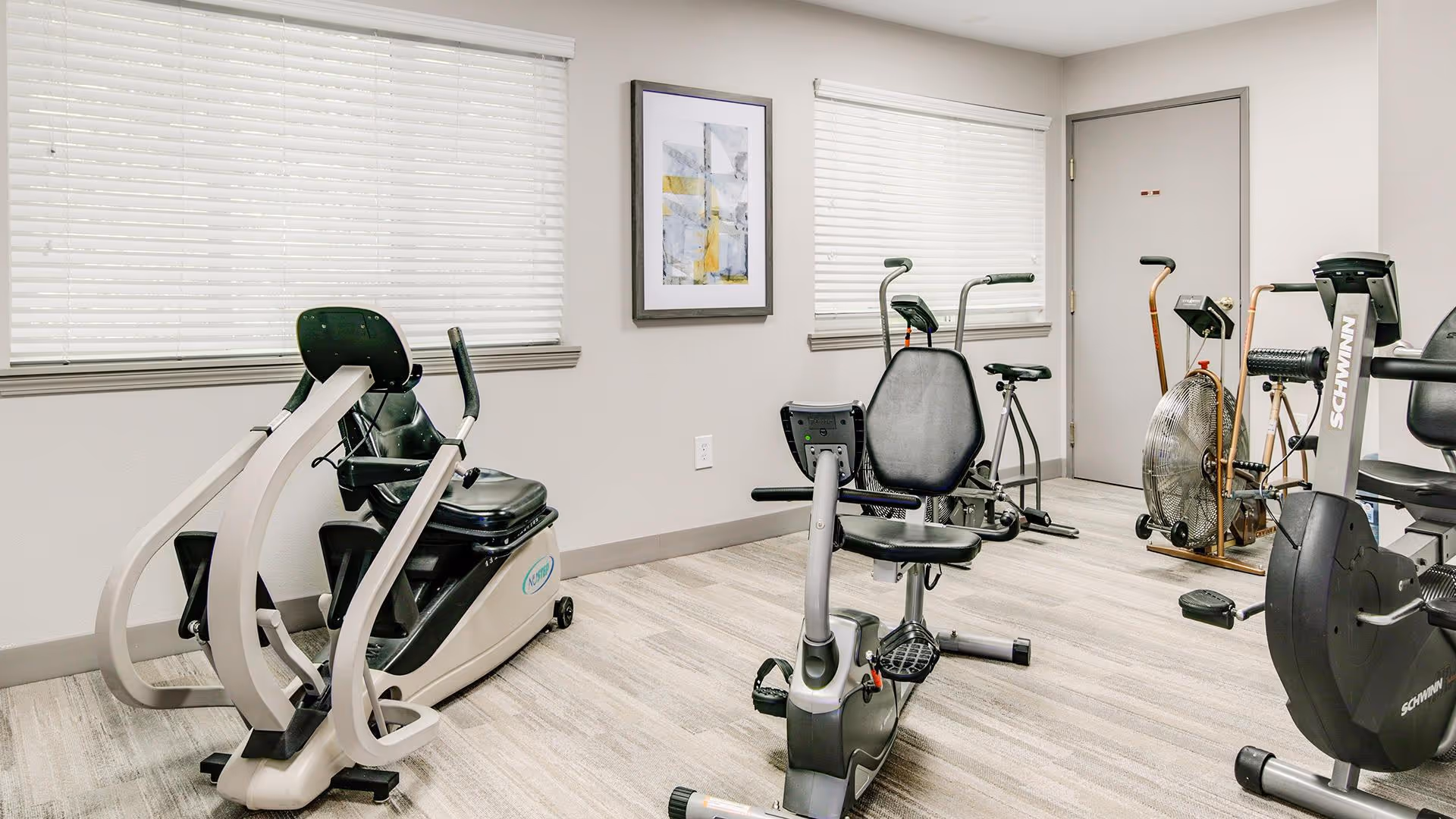 A small fitness room with several exercise machines including recumbent bikes and an air resistance bike. The room has light-colored walls, two windows with white blinds, a framed abstract painting on the wall, and a closed door.