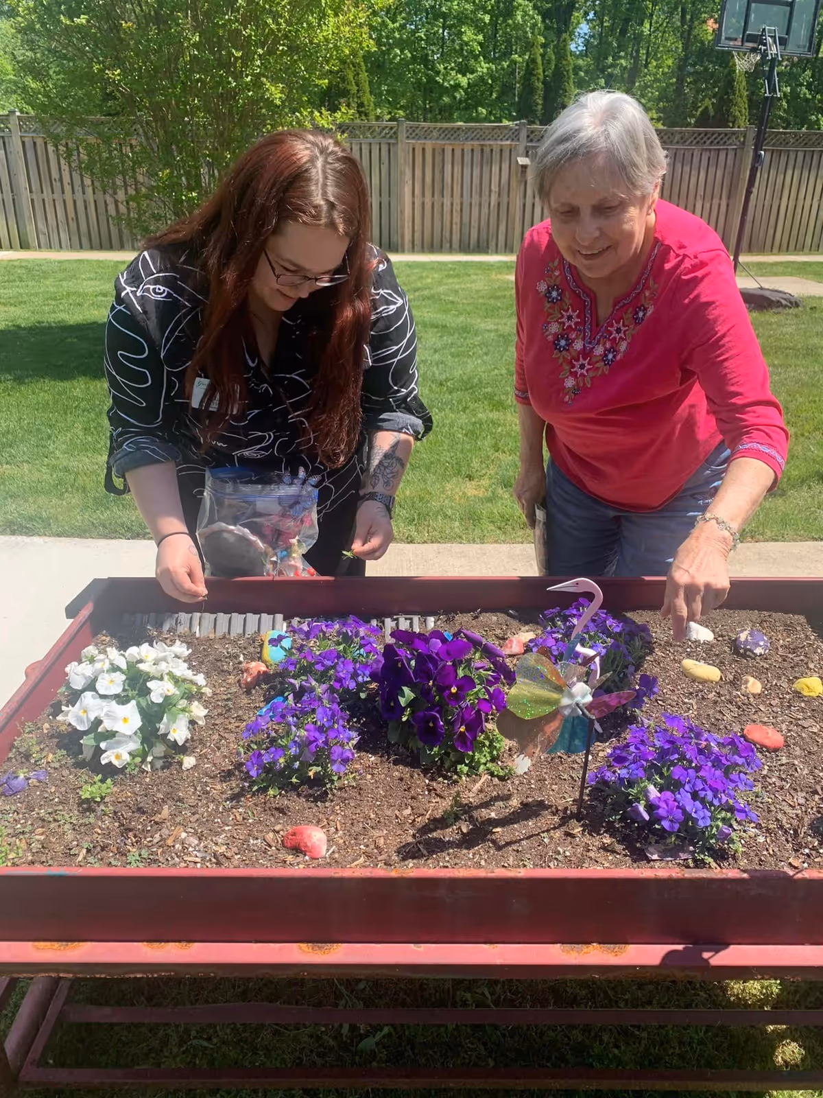 Two women tending to a raised garden bed outdoors on a sunny day. The garden bed contains purple and white flowers, decorative painted rocks, and a small pinwheel. One woman with long brown hair and glasses is holding a plastic bag, while the older woman with short gray hair and a pink embroidered top is pointing at something in the garden bed. A wooden fence and green trees are visible in the background.