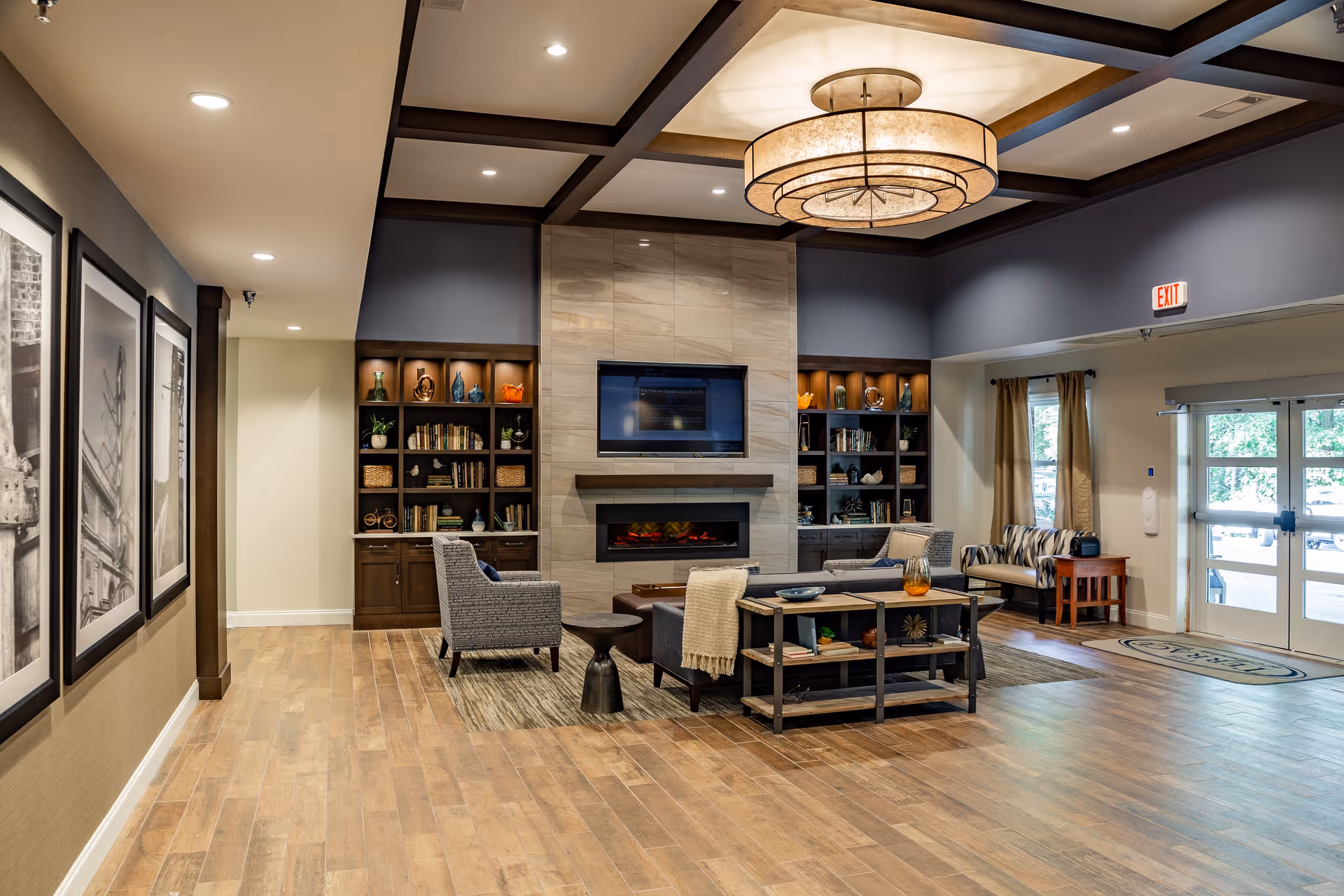 A spacious and well-lit senior living facility common area featuring a modern fireplace with a mounted TV above it, flanked by built-in shelves filled with books and decorative items. The seating area includes comfortable chairs and a sofa arranged around a coffee table. The room has wooden flooring, recessed lighting, and large windows near the entrance door allowing natural light to enter.