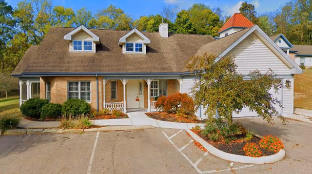 Front exterior view of a single-story assisted living facility building with a brown shingle roof, two dormer windows, a covered porch with white railings, and a small landscaped area with bushes and flowers near the entrance. The parking area is visible in front of the building, surrounded by trees with green foliage in the background.