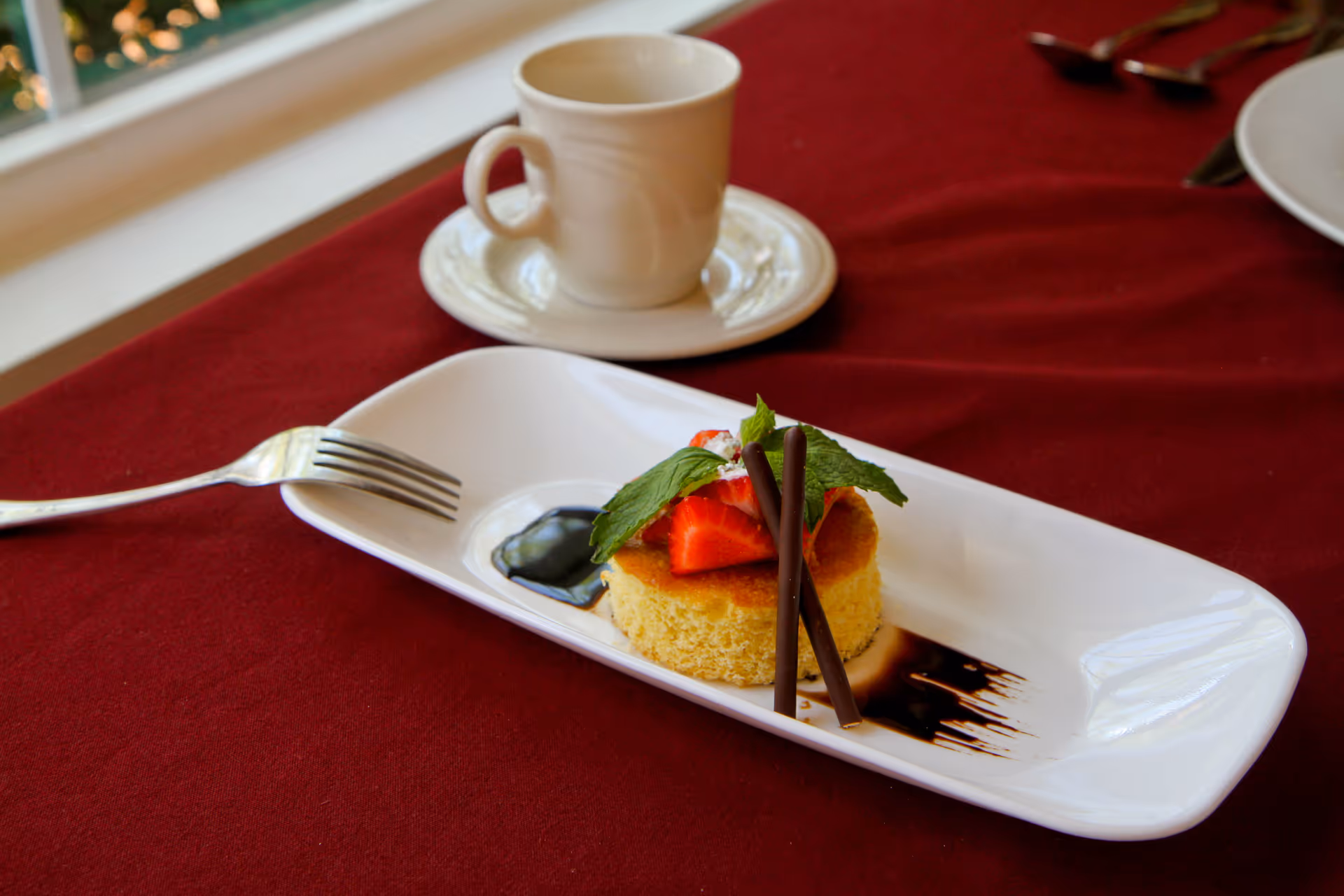 Plated dessert topped with strawberries, mint, and chocolate sticks on a white rectangular plate on a red tablecloth with a coffee cup in the background.