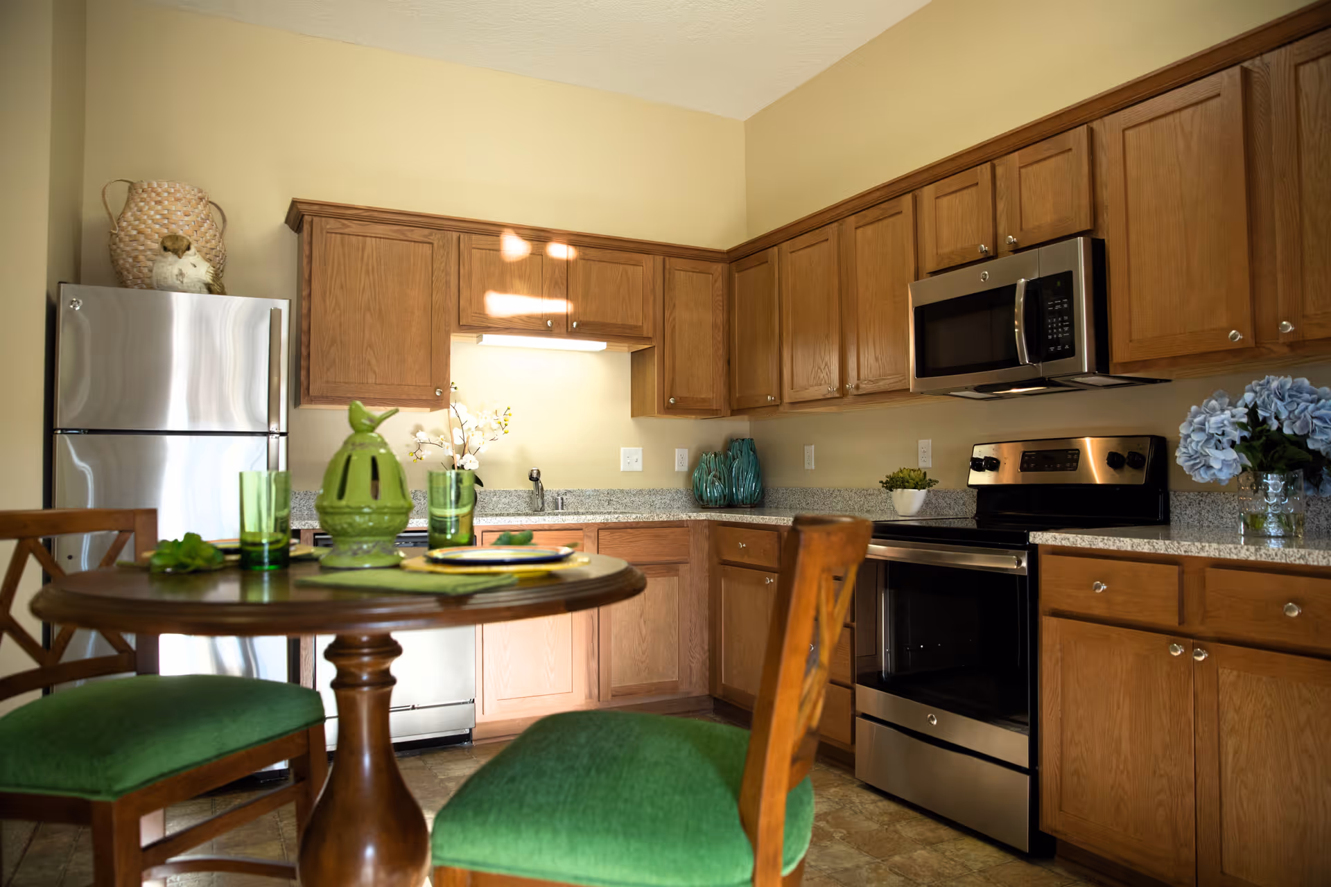 A kitchen with wooden cabinets, stainless steel refrigerator, oven, and microwave. There is a round wooden table with green cushioned chairs and green tableware. Decorative items include a green ceramic bird, flowers in vases, and a small plant on the countertop.