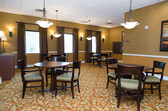A senior living facility dining room with round wooden tables and cushioned chairs arranged on a patterned carpet. The room has beige walls with white trim, three windows with brown curtains, and hanging light fixtures providing warm lighting.
