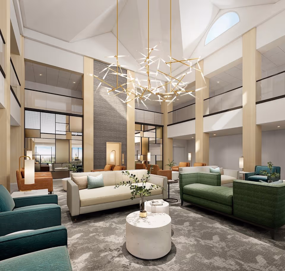 A spacious and modern senior living facility lounge area with high ceilings and large windows allowing natural light. The room features multiple comfortable sofas and armchairs in green, beige, and brown tones arranged around small round tables with decorative plants. A contemporary chandelier with multiple light rods hangs from the ceiling, and the walls have a mix of light wood paneling and textured surfaces.