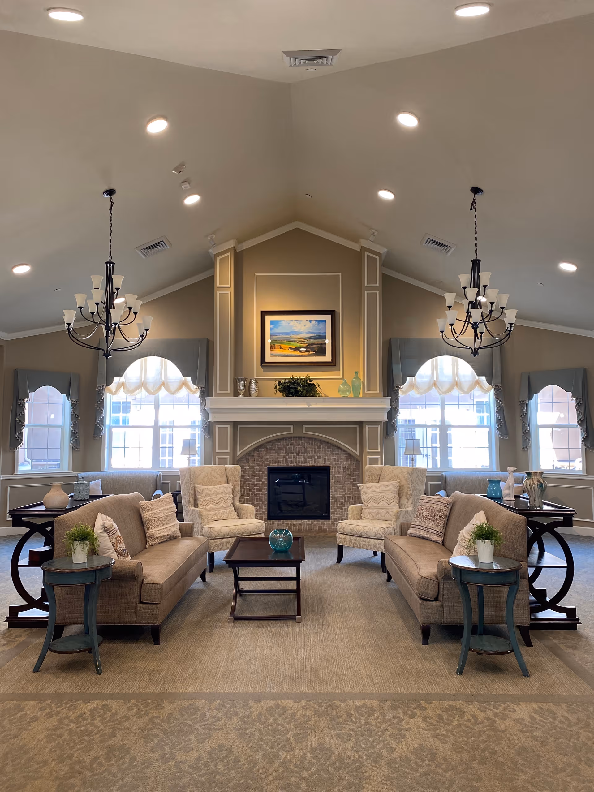 A spacious and elegant living room with two beige sofas facing each other, two patterned armchairs, a central coffee table, and side tables with plants. The room features a fireplace with a framed landscape painting above it, large windows with decorative curtains, and two black chandeliers hanging from a vaulted ceiling with recessed lighting.