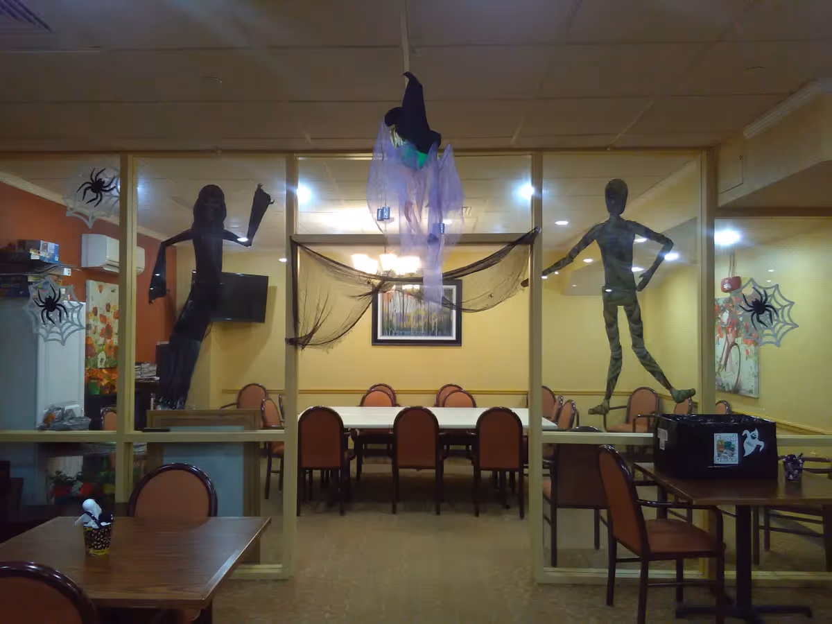 Interior view of a dining area decorated for Halloween with hanging ghost and witch figures, spider web decals on glass walls, and tables with chairs arranged around them.