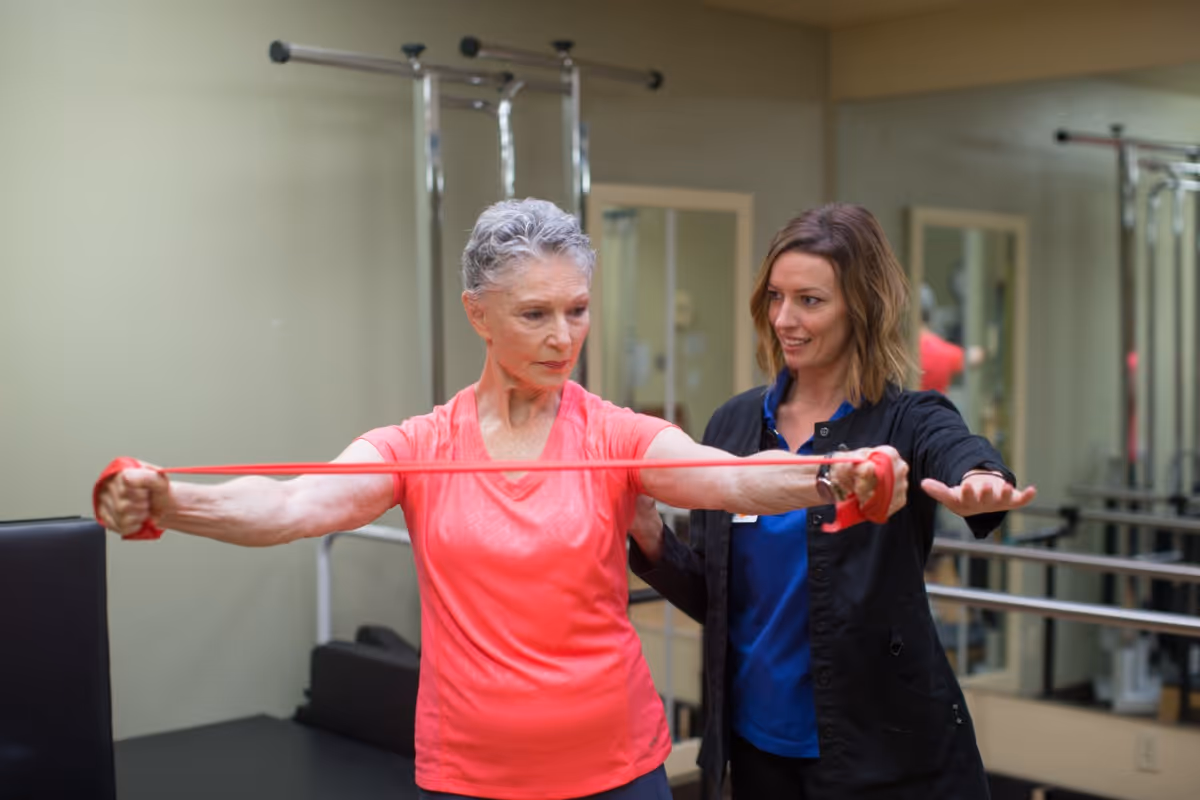 An elderly woman in a pink shirt is exercising with a red resistance band while a younger female instructor in a black jacket and blue shirt assists her in a fitness or therapy room with mirrors and exercise equipment.