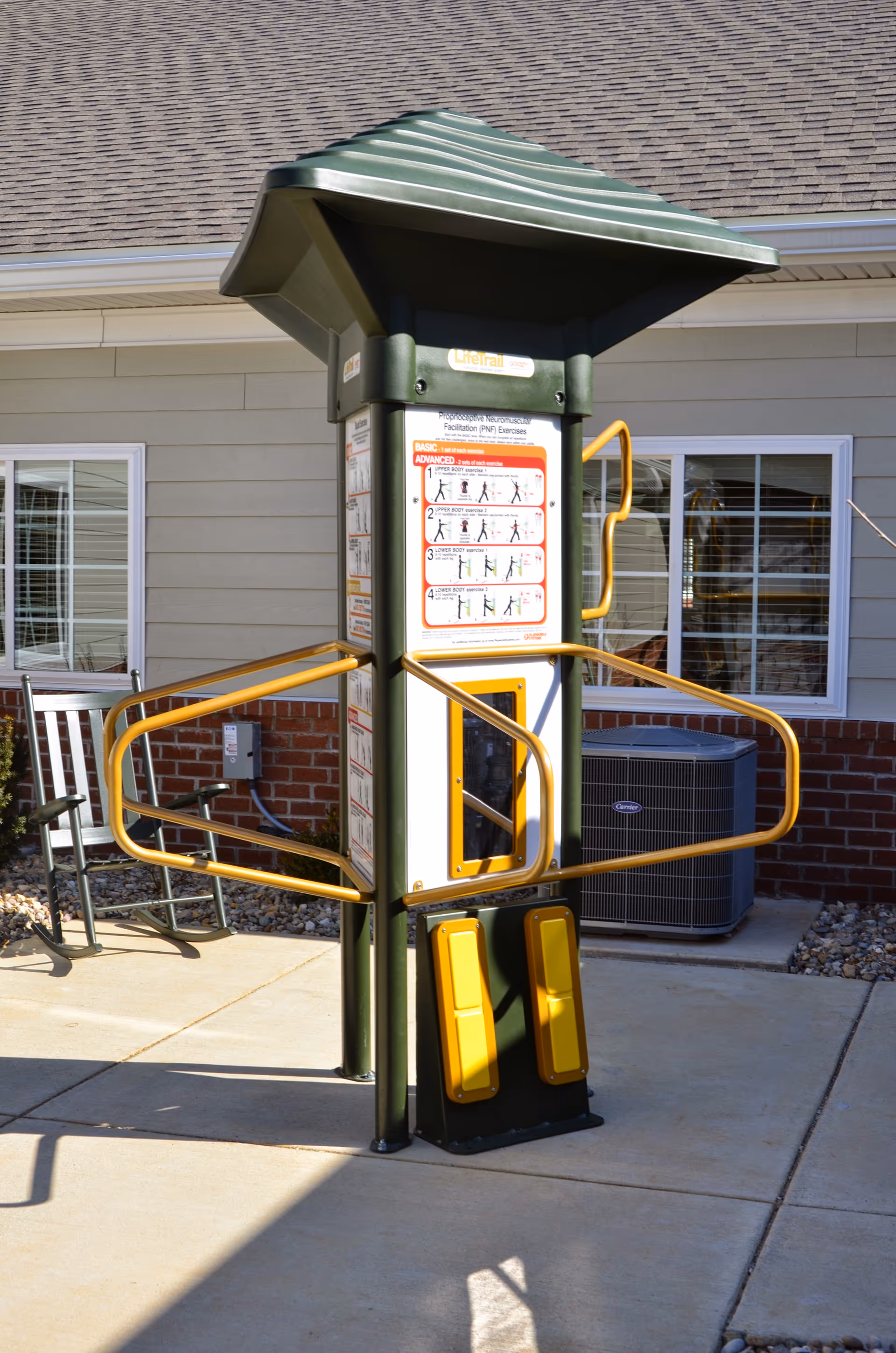 Outdoor exercise equipment with instructional signs, yellow handrails, and foot pedals, located on a concrete patio in front of a building with windows and a brick lower wall.