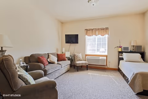 Bright studio-style room with a sofa, armchair, wall-mounted TV and a single bed beside a window and air conditioner.