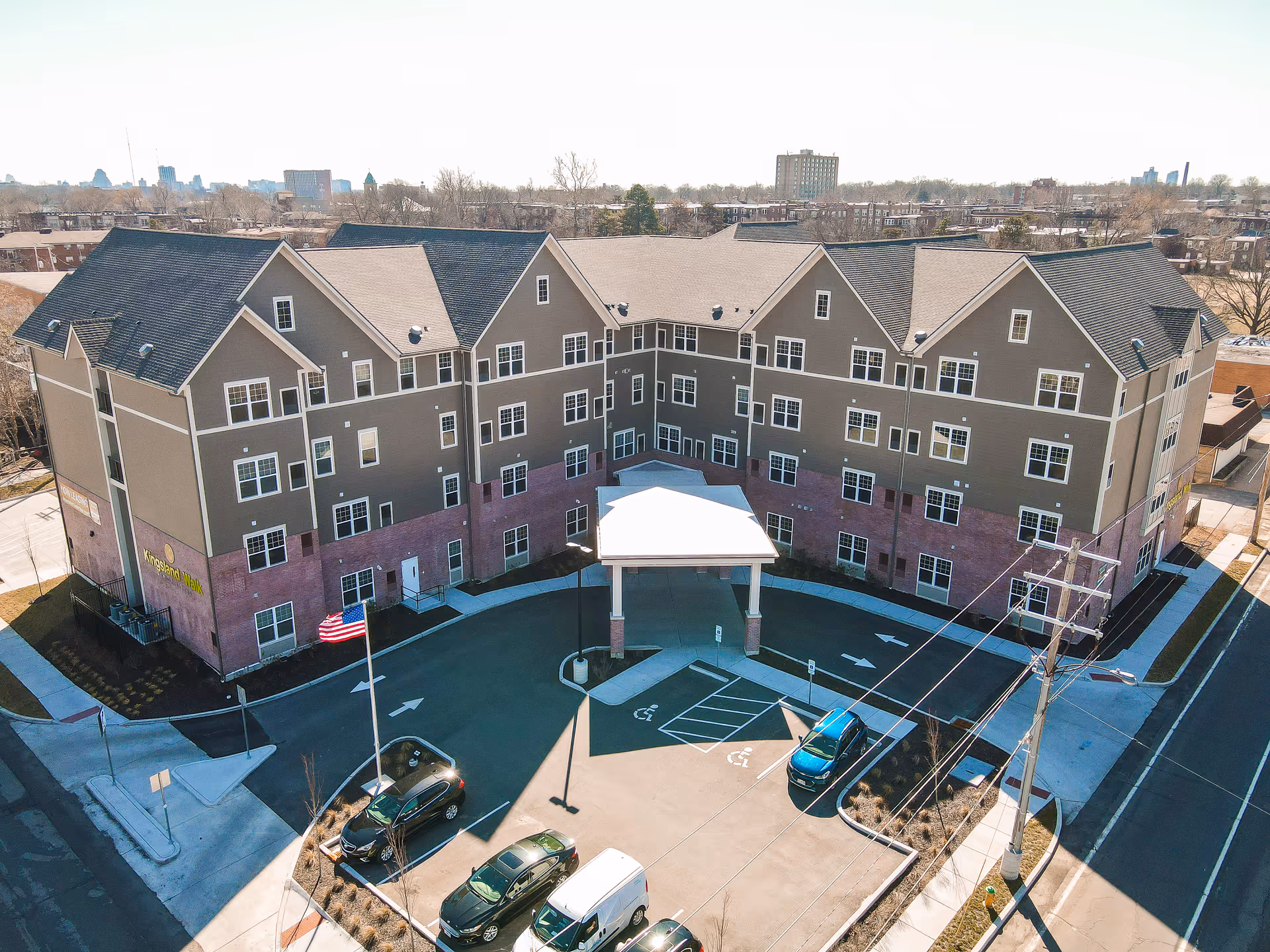 Aerial view of a multi-story senior living building with a covered entrance, parking lot, and an American flag.