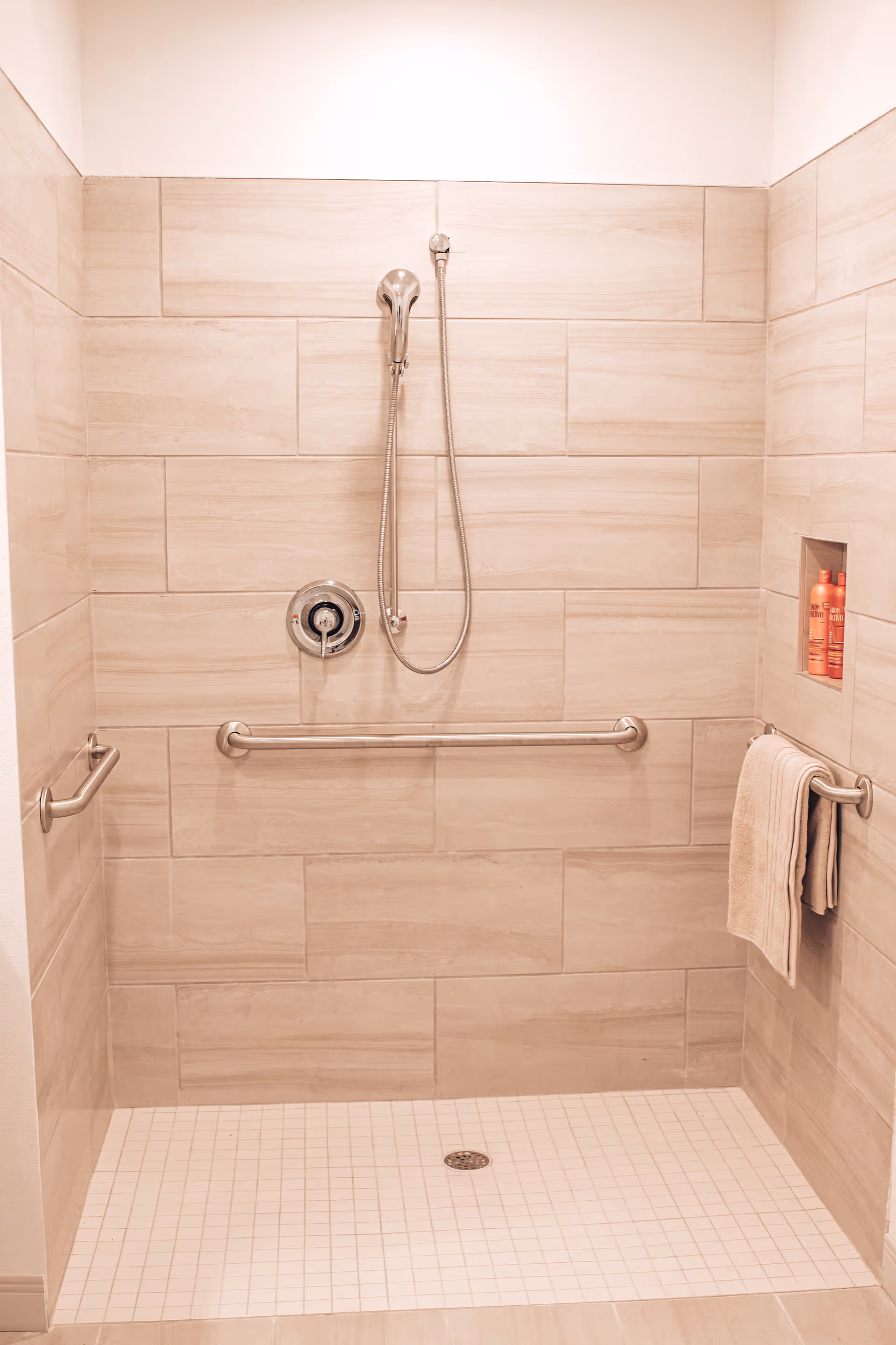 Tiled walk-in shower with a handheld showerhead, horizontal grab bars, a recessed niche with bottles, and a towel on a rail.