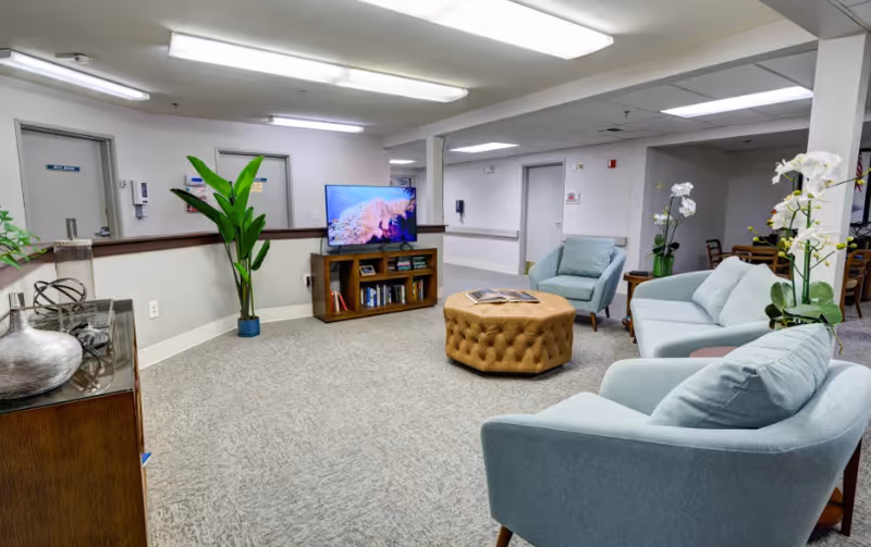 A bright senior living common room with light blue armchairs, a tufted ottoman, a TV on a wooden console, and potted plants.
