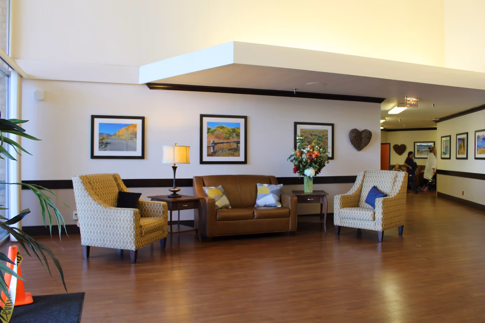 A cozy seating area in a senior living facility with two patterned armchairs and a brown leather sofa arranged around two wooden side tables. One table has a lamp, and the other has a vase with a colorful flower arrangement. The walls are decorated with framed landscape photographs and a heart-shaped wall decoration. The floor is wooden, and there is a hallway visible in the background with a few people.