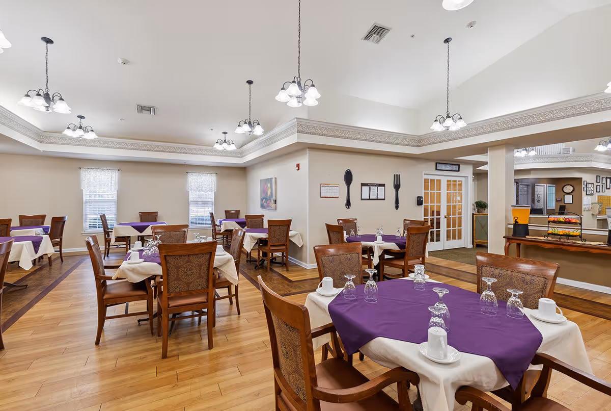 A dining room with several wooden tables and chairs arranged neatly. Each table is covered with a white tablecloth and a purple table runner, set with upside-down glasses and white coffee cups. The room has wooden flooring, beige walls, and multiple ceiling light fixtures. There are two windows with white curtains, wall decorations including large fork and spoon wall art, and a buffet area with a beverage dispenser and fruit stand.