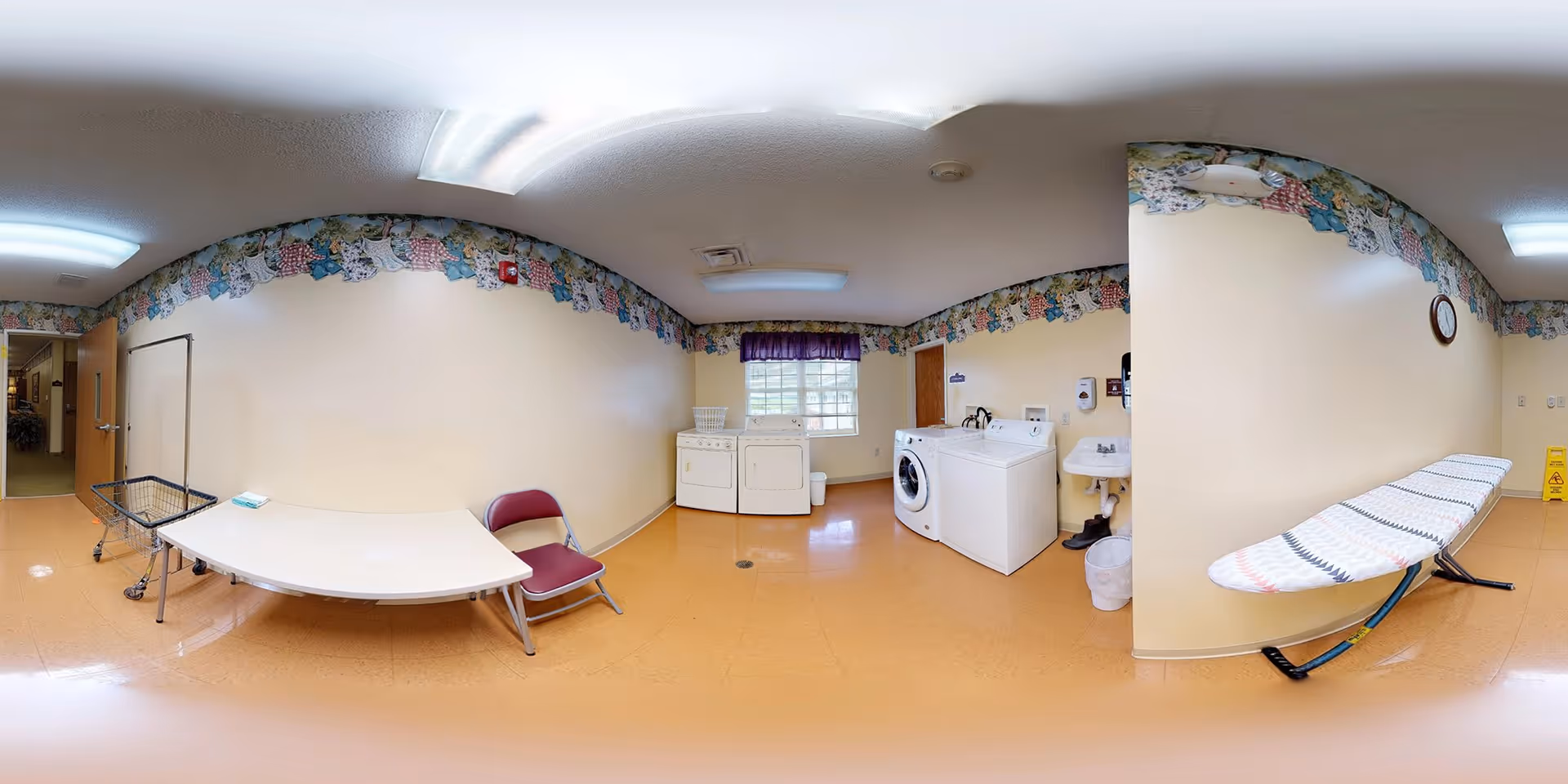 A laundry room with a washing machine, dryer, ironing board, folding table, and a chair. The room has beige walls with a floral border near the ceiling, a window with a purple valance, and a small sink. The floor is a light brown color, and there is a laundry cart and a caution wet floor sign visible.
