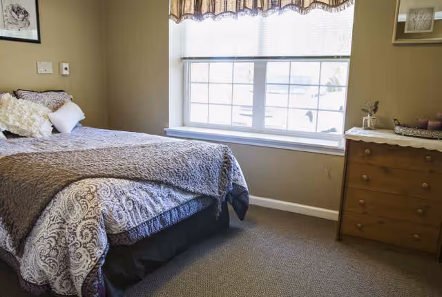 A cozy bedroom with a bed covered in patterned bedding and a gray throw blanket. There are pillows on the bed, a window with blinds and a valance letting in natural light, and a wooden dresser with a lace runner and decorative items on top.