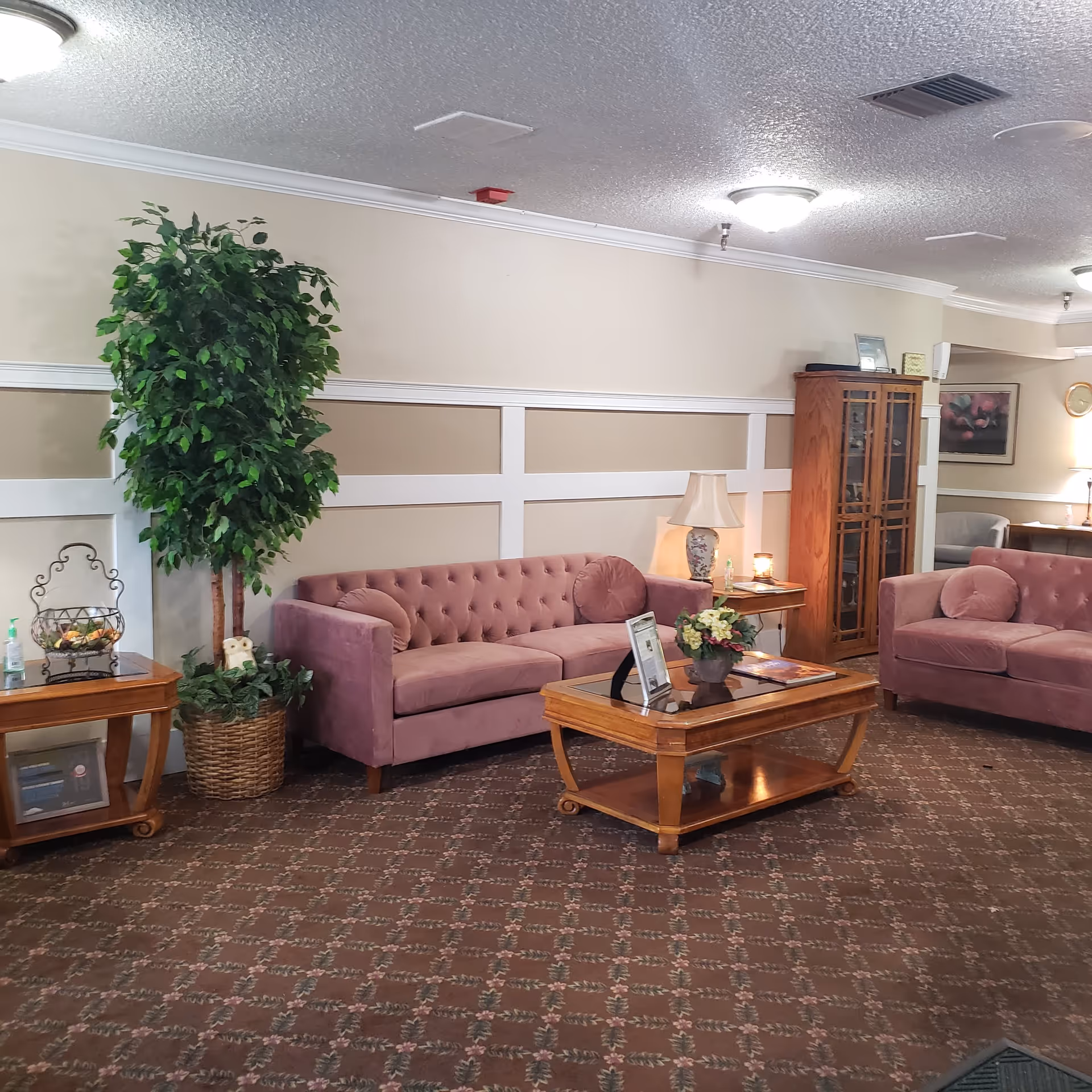 A cozy living room area with two mauve tufted sofas, a wooden coffee table with a glass top and floral centerpiece, a wooden side table with a lamp, a tall potted plant, and a wooden cabinet with glass doors. The room has beige walls with white trim and patterned carpet flooring.