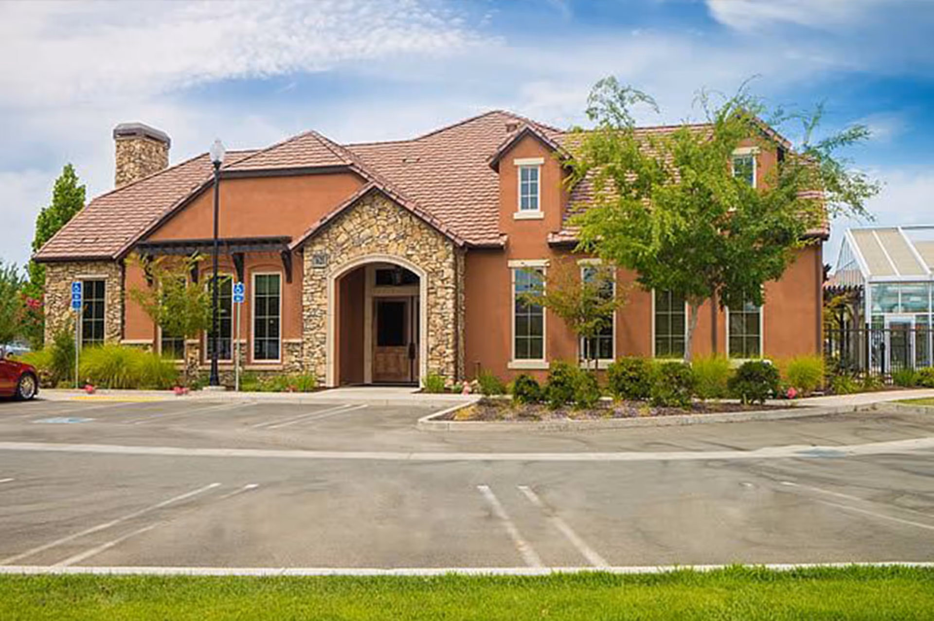 Two-story stone-and-stucco senior living building with an arched entrance, landscaping, and a parking lot in front.