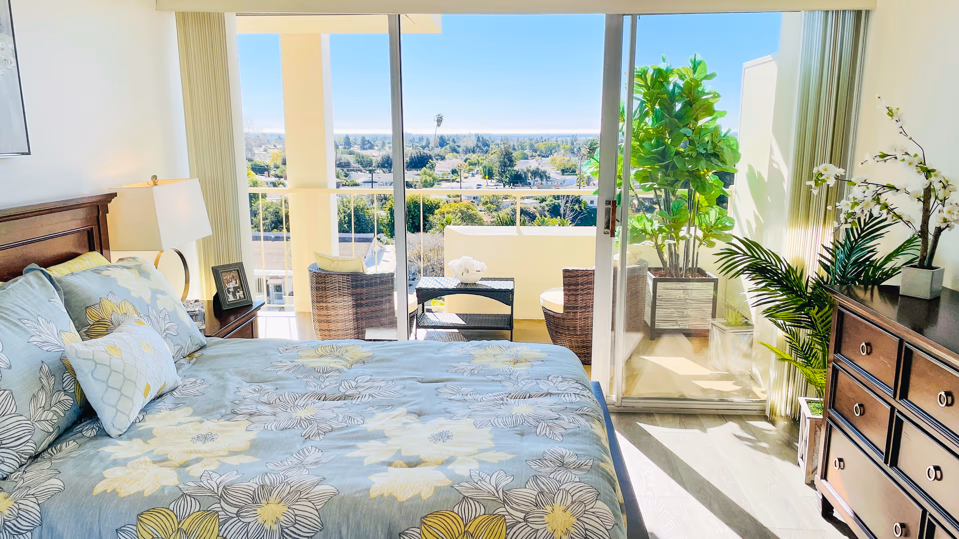 Bright bedroom with a bed covered in floral bedding, a wooden nightstand with a lamp and picture frame, a wooden dresser with decorative plants, and a sliding glass door leading to a balcony with two wicker chairs and a small table overlooking a scenic view.