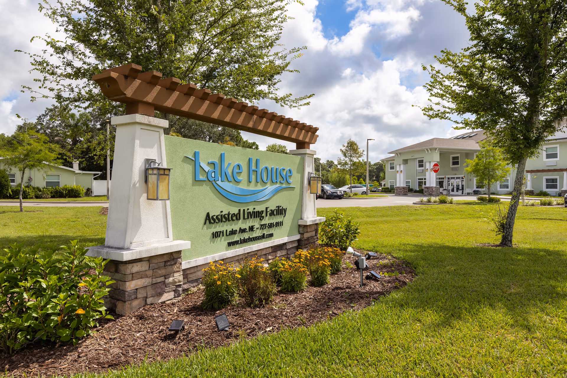 Outdoor view of the entrance sign for Lake House Assisted Living Facility with the building and parking lot visible in the background under a partly cloudy sky.