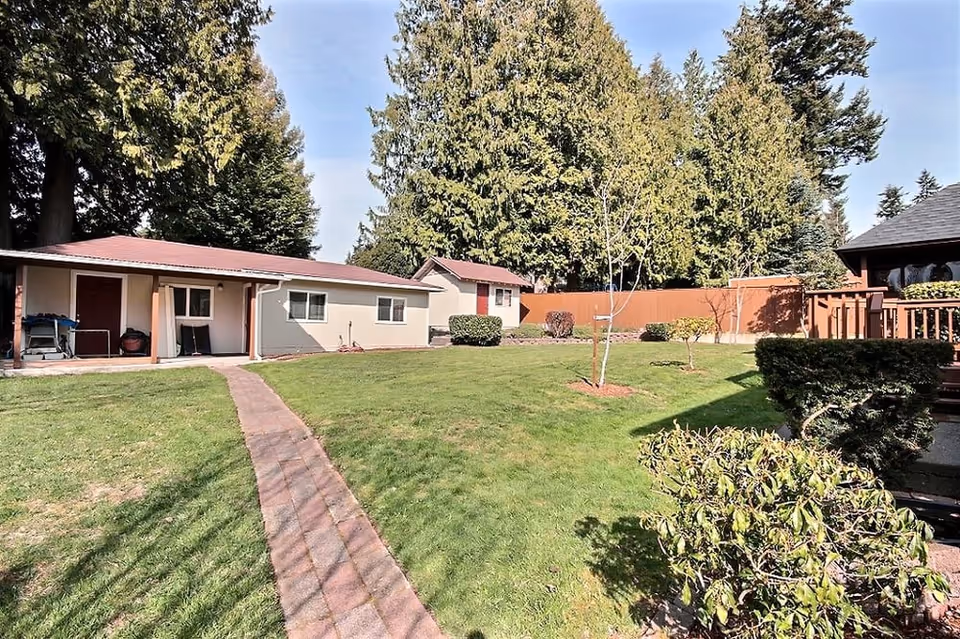 A well-maintained outdoor area with a green lawn, a paved walkway, several small trees and bushes, and multiple single-story buildings with red roofs surrounded by tall evergreen trees under a clear sky.